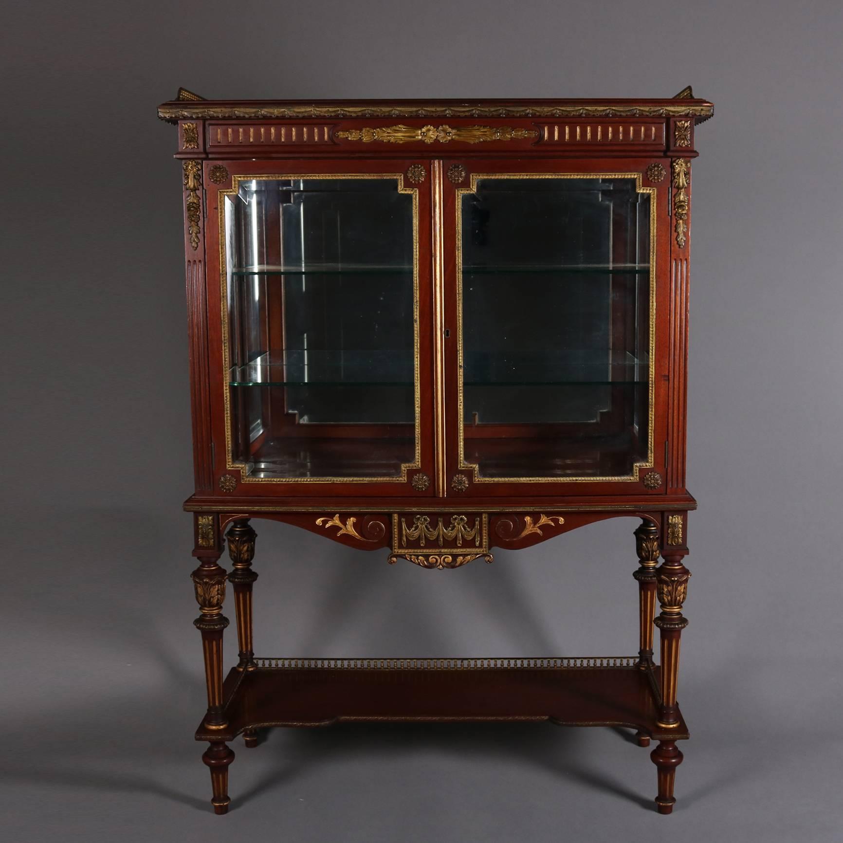 Antique French Louis XVI style vitrine features mahogany construction, cast bronze accoutrements and upper gallery, gilt highlights, swag and foliate decorated apron, two glass front doors opening to mirrored back interior with glass shelving, lower