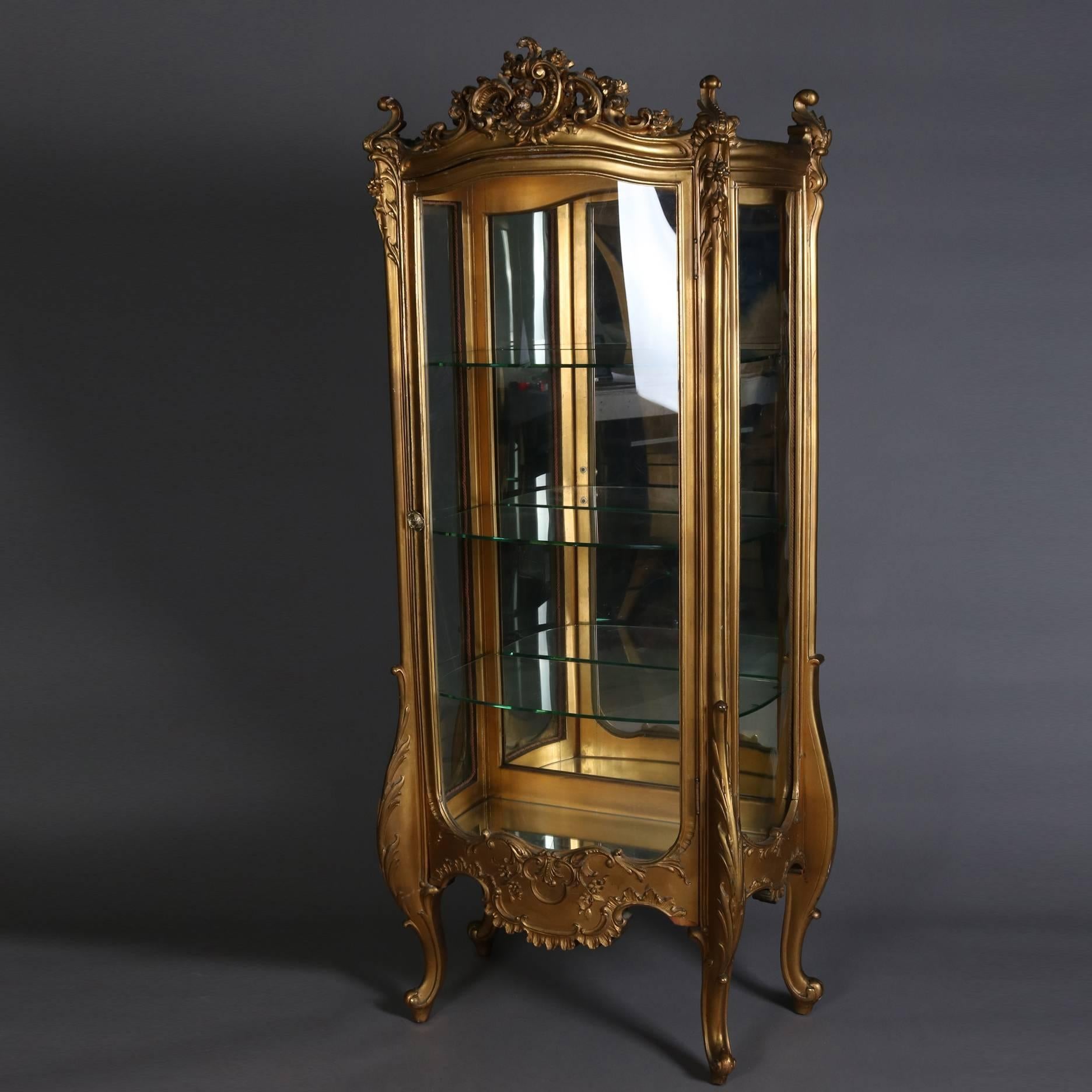 Antique French Louis XIV vitrine features giltwood case with scroll, foliate and floral decoration, pierced crest with cartouche, seated on exaggerated cabriole legs, single bow front glass door opens to mirror back interior display with glass