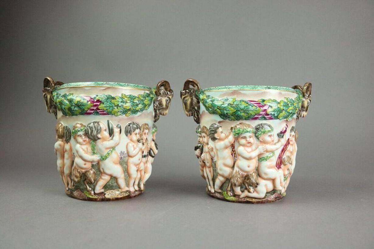 Pair of figural Capodimonte open urns or cups feature hand-painted Cupids (Putti), ram handles, and crown N mark, late 19th century.