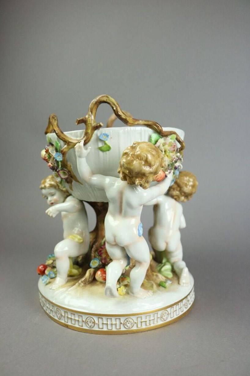 German figural Schierholz Meissen style porcelain compote features hand-painted putti (Cupids) lifting basket with foliate accents, and early blue underglaze mark of crown and shield with oak leaves on base, circa 1900.

C.G. Schierholz (&