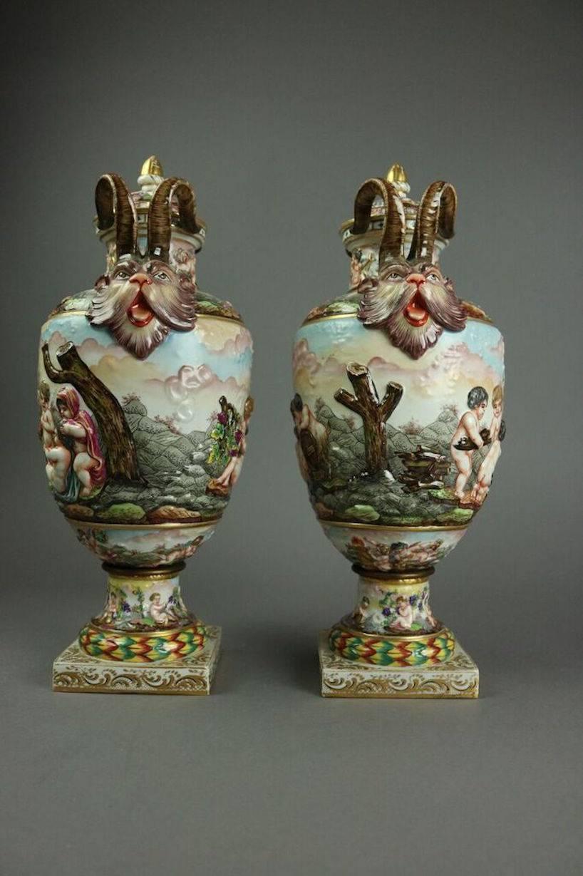 Pair of classically shaped Italian Capodimonte figural covered urns feature hand-painted scene of continuous cupid (putti) procession, handles with figurehead terminations, and blue crown N marks, 19th century. 

