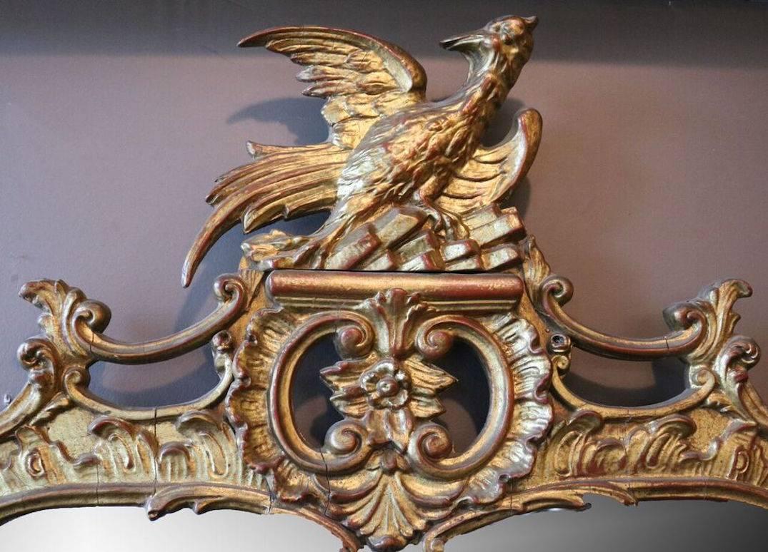 A George III style carved giltwood surround of S and  C-scrolls, foliate and floral pendants with a pierced crest centering a dimensionally carved feathered phoenix in heraldry and a rectangular mirror panel, circa 1940. Measures: 50"H.