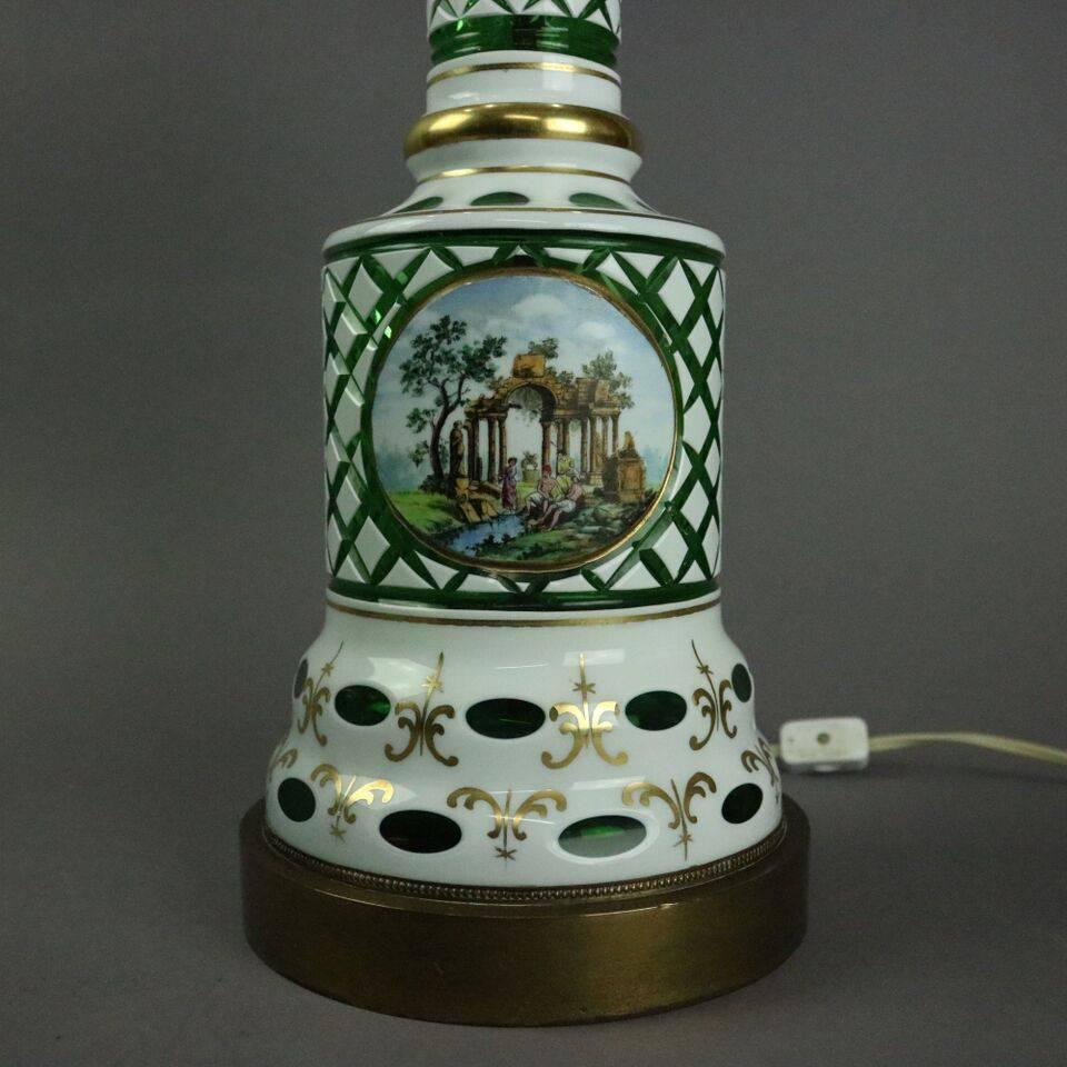 Monumental antique Bohemian style table lamp features Opaline overlay cut to emerald green glass in diamond and thumb print patterns with hand-painted Persian river scene reserve and floral band on column with gilt highlights topped by molded thumb