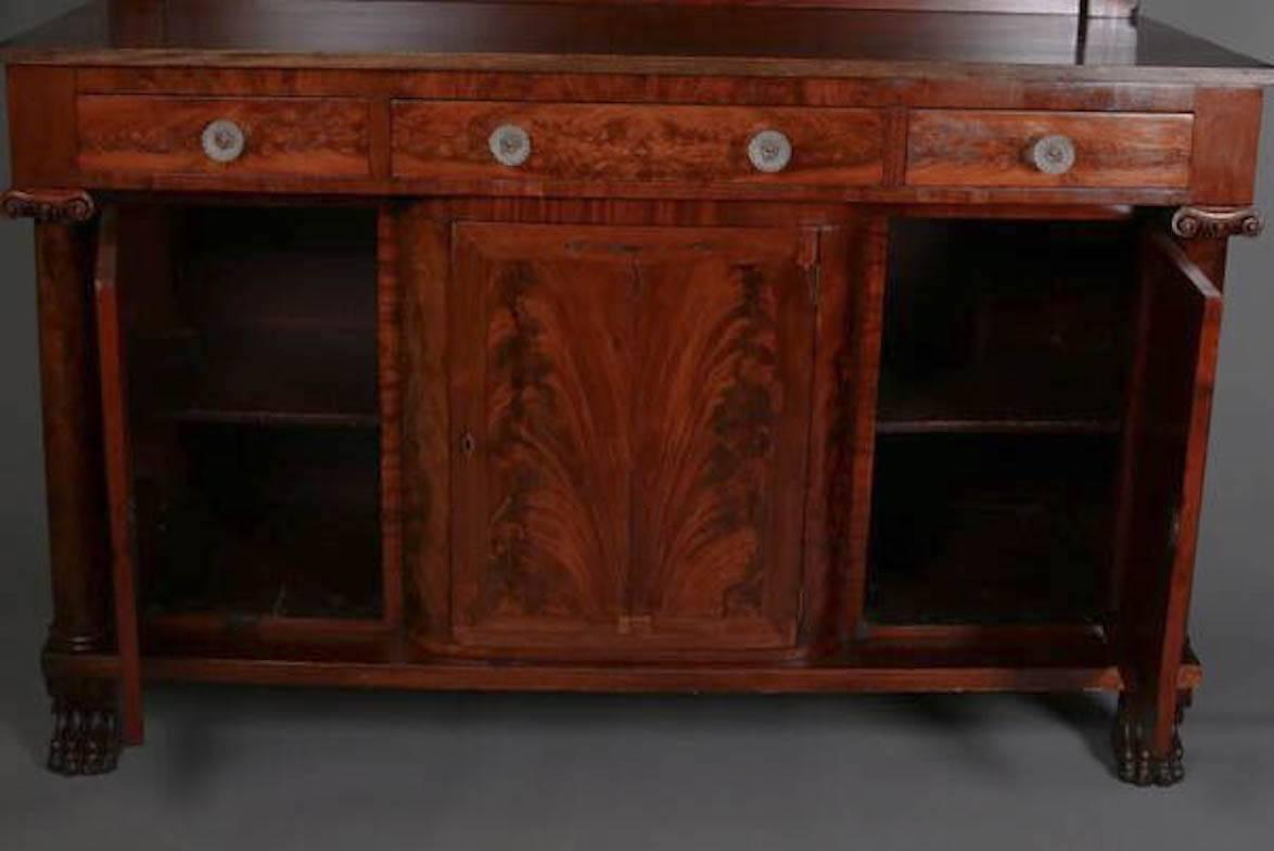 Antique Period American Empire sideboard features serving and display surface with backsplash above three drawers and three lower cabinet compartments with bookmatched flame mahogany, flanked by Corinthian form columns with carved capitals, seated