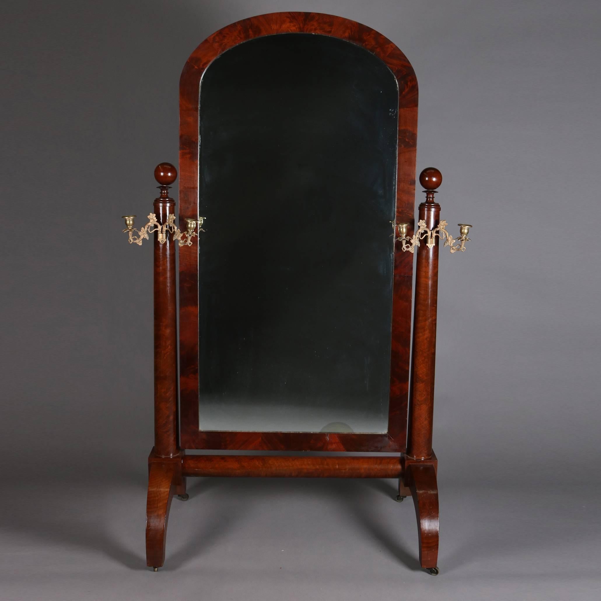 Antique American Empire Quervelle school cheval dressing mirror features flame mahogany frame, arched mirror flanked by grape and leaf form double candelabra, and seated on trestle base with arched legs each having central cast bronze medallion,