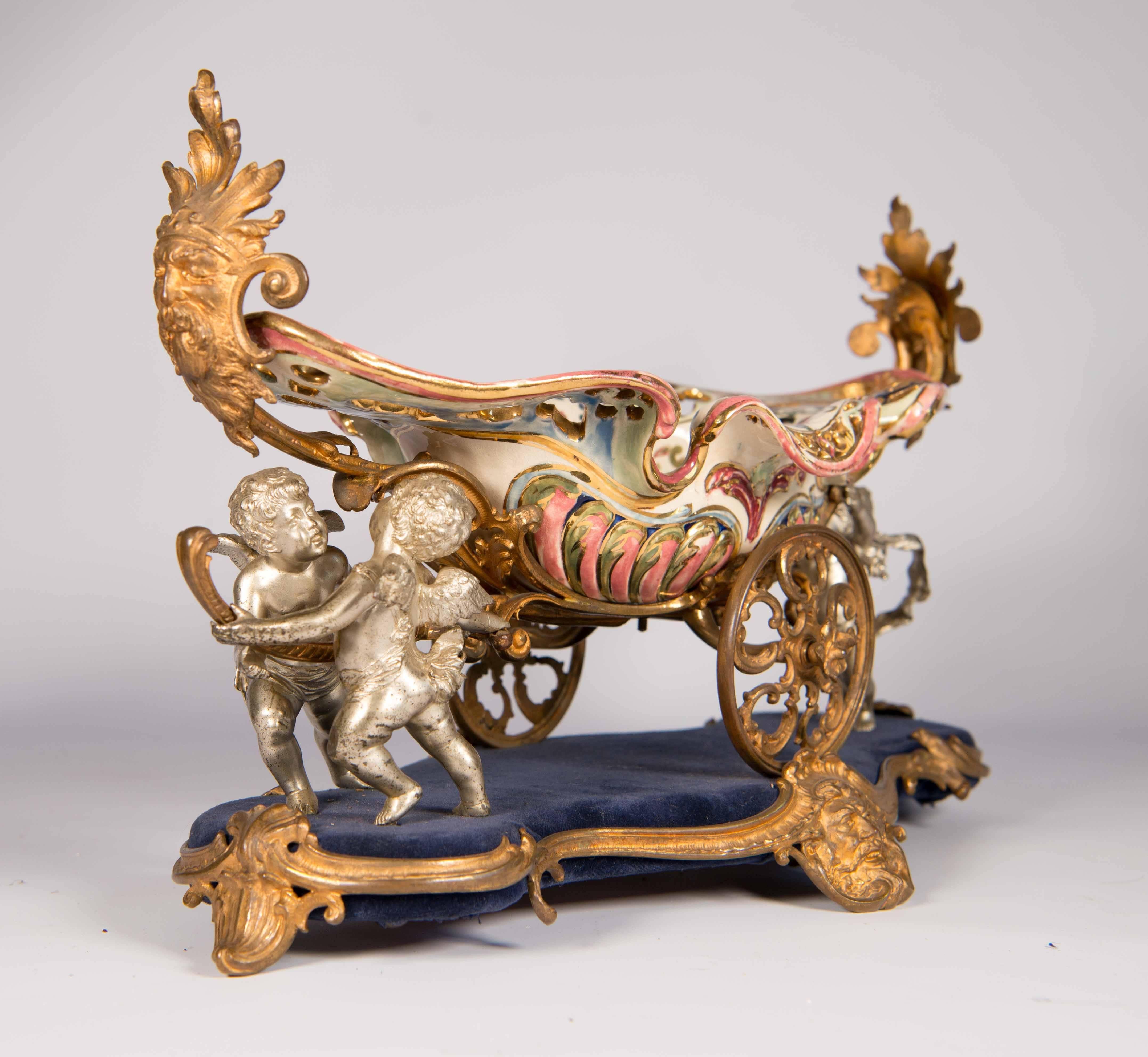 Porcelain and bronze combination table centrepiece. The item was made in Budapest, circa 1920 and holds the signature Fischer J. Budapest. The centrepiece has three silver plated cherubs pushing a porcelain basket on wheels. Everything is in mint