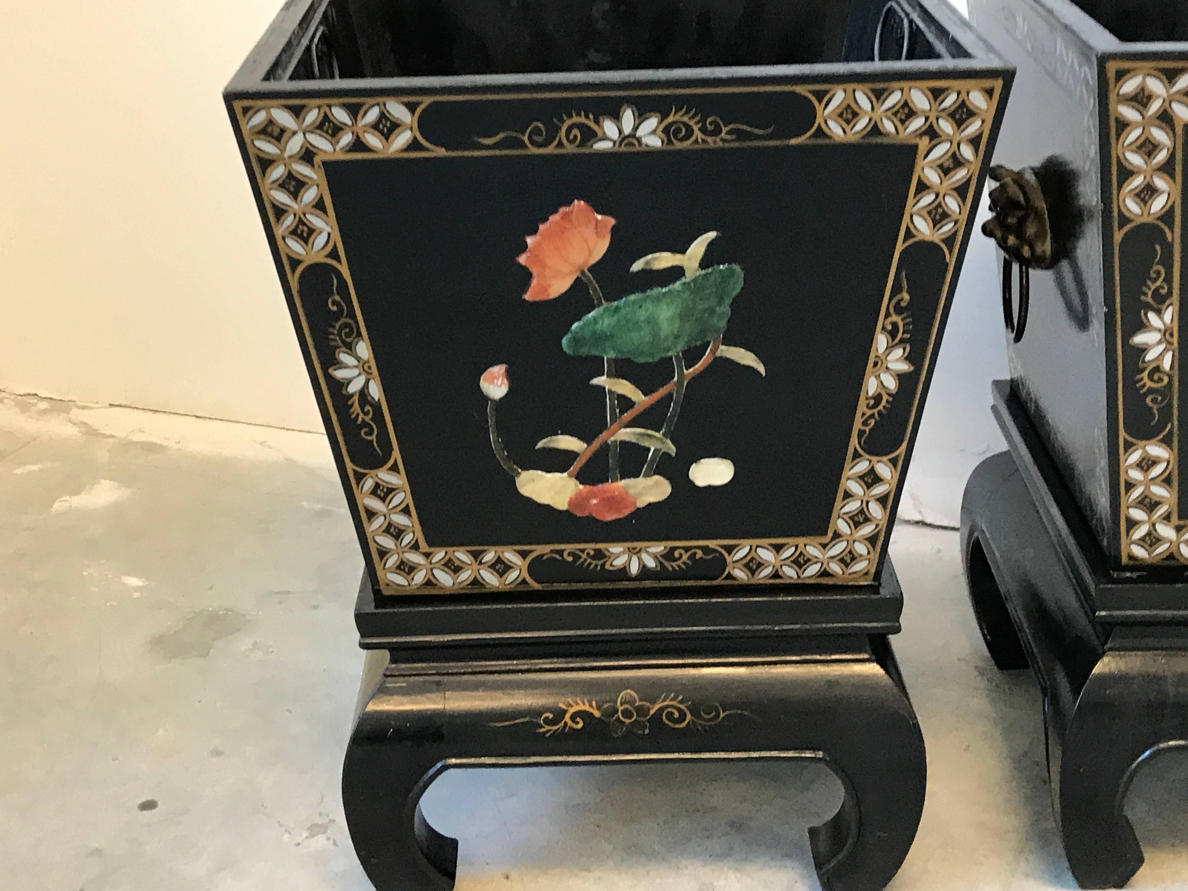Offered is a gorgeous, pair of 1960s, black and gold Asian planters. Each planter rests on a Ming style stand. Floral motif with stone inlay, hand-painted fretwork border and brass foo dog handles on each. Each includes metal liner.