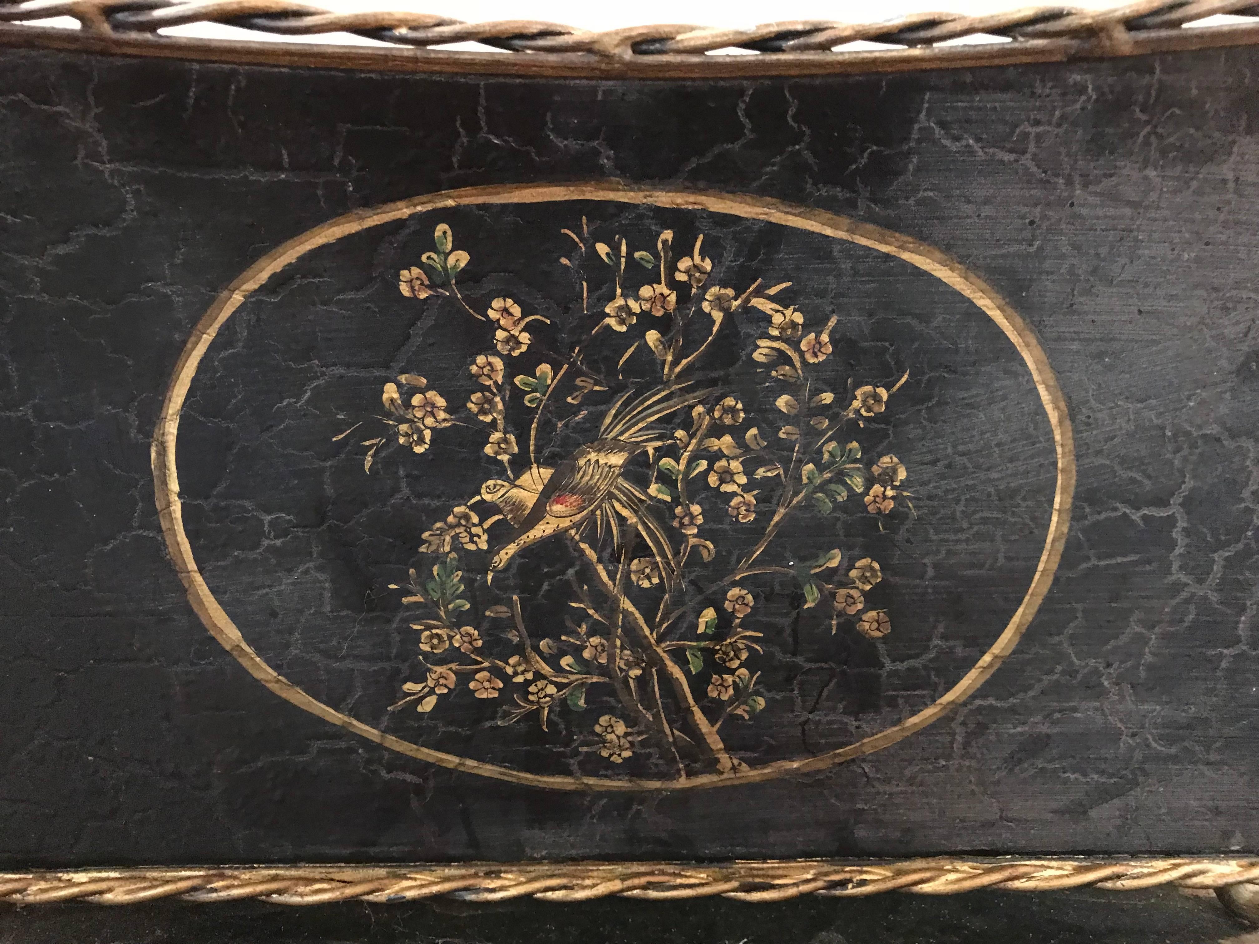 Offered is a beautiful, 1970s black and gold tole cachepot planter. The piece has a gorgeous, hand-painted chinoiserie floral and bird motif, one on each of the longer sides. Gold-painted braiding along the top and bottom. Brass lion head handles,