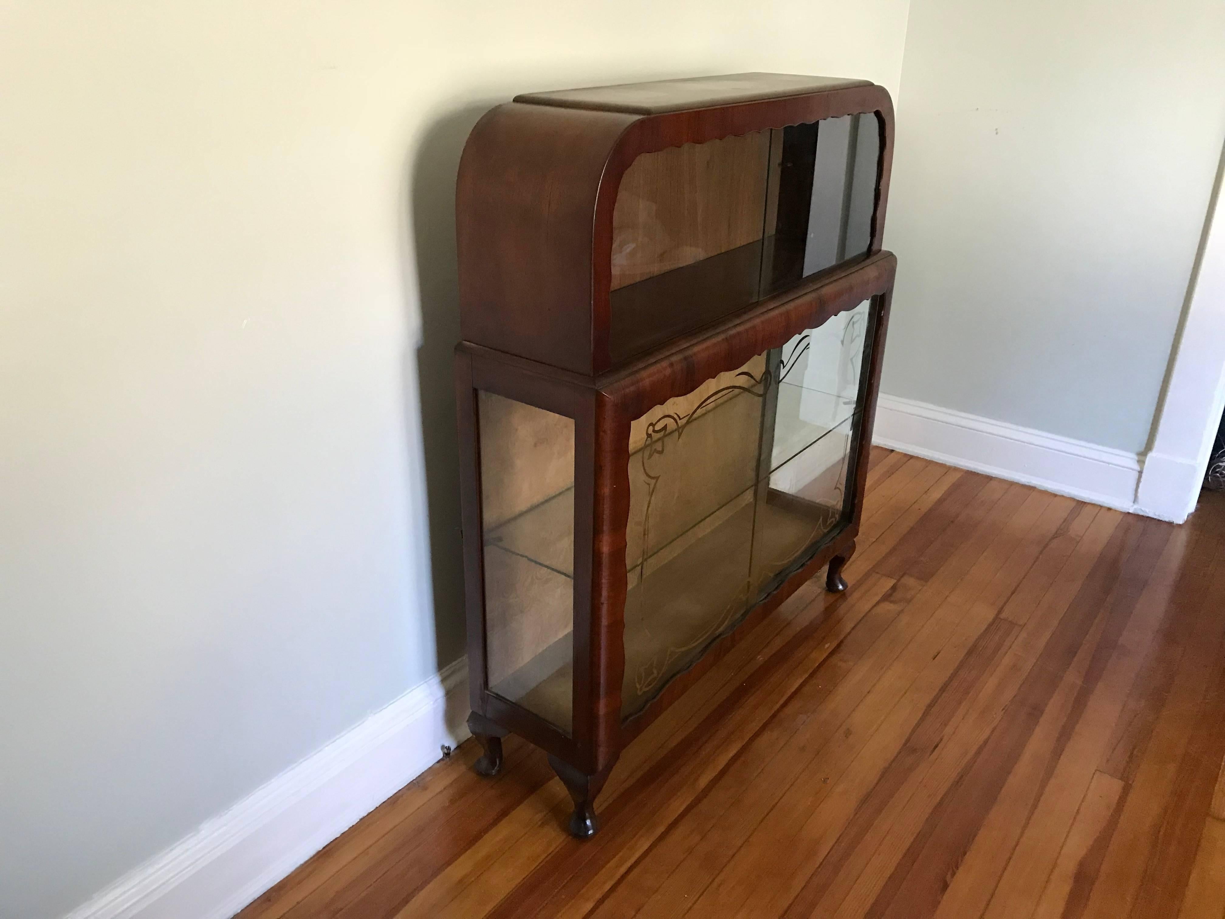 Offered is a gorgeous, 1920s French Art Deco glass-front curio display cabinet. The piece has a stunning, scalloped woodwork detailing, framing the sliding glass doors. The doors have a beautiful, silver-leaf stencil work around the edges.