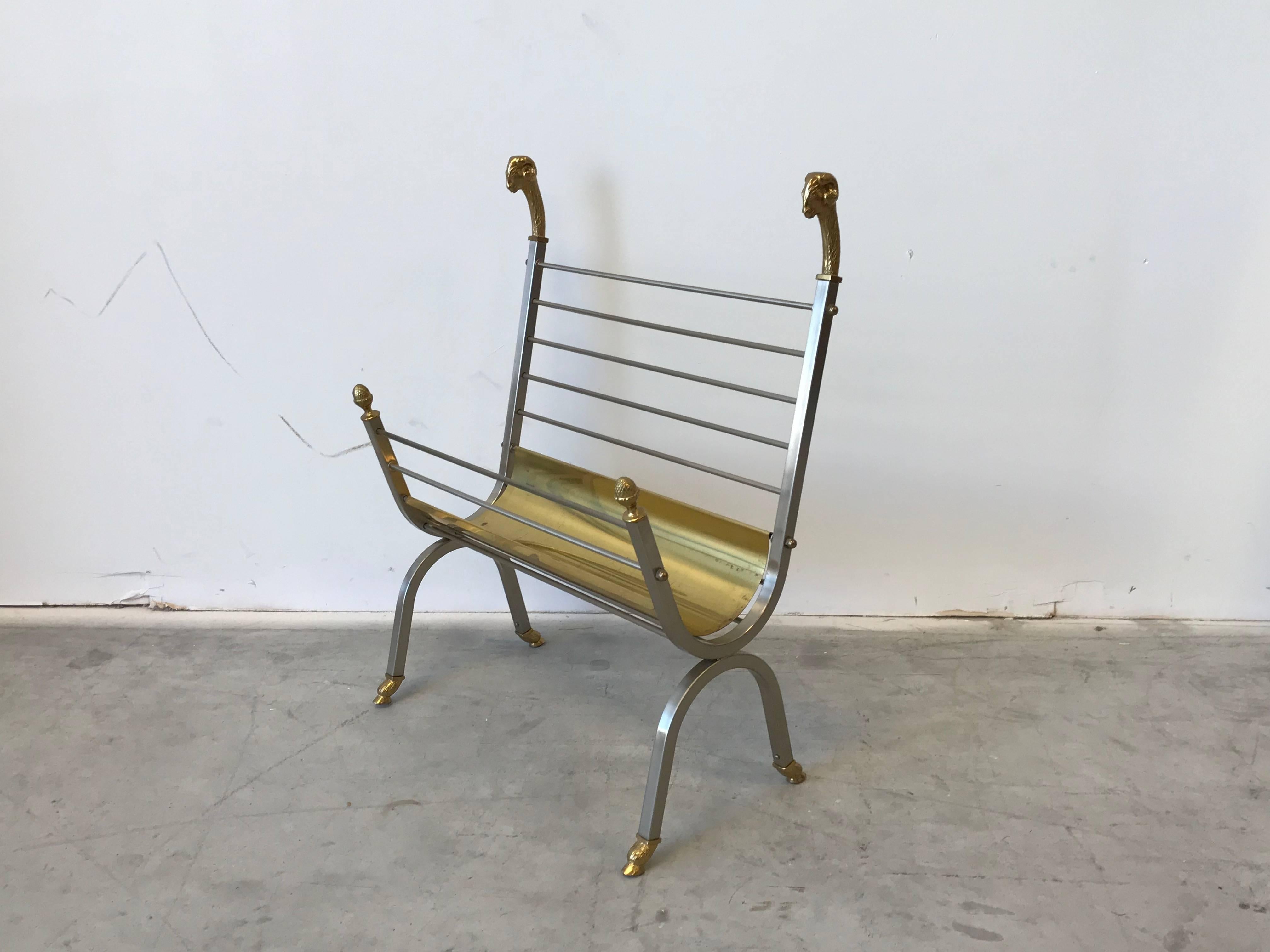 Offered is a stunning, 1970s Italian Maison Jansen style, brass and steel magazine rack. The elegant sides are adorned with brass rams heads and pineapple/acorn finials. The piece is raised, with brass hoof-feet. The piece includes a removable,