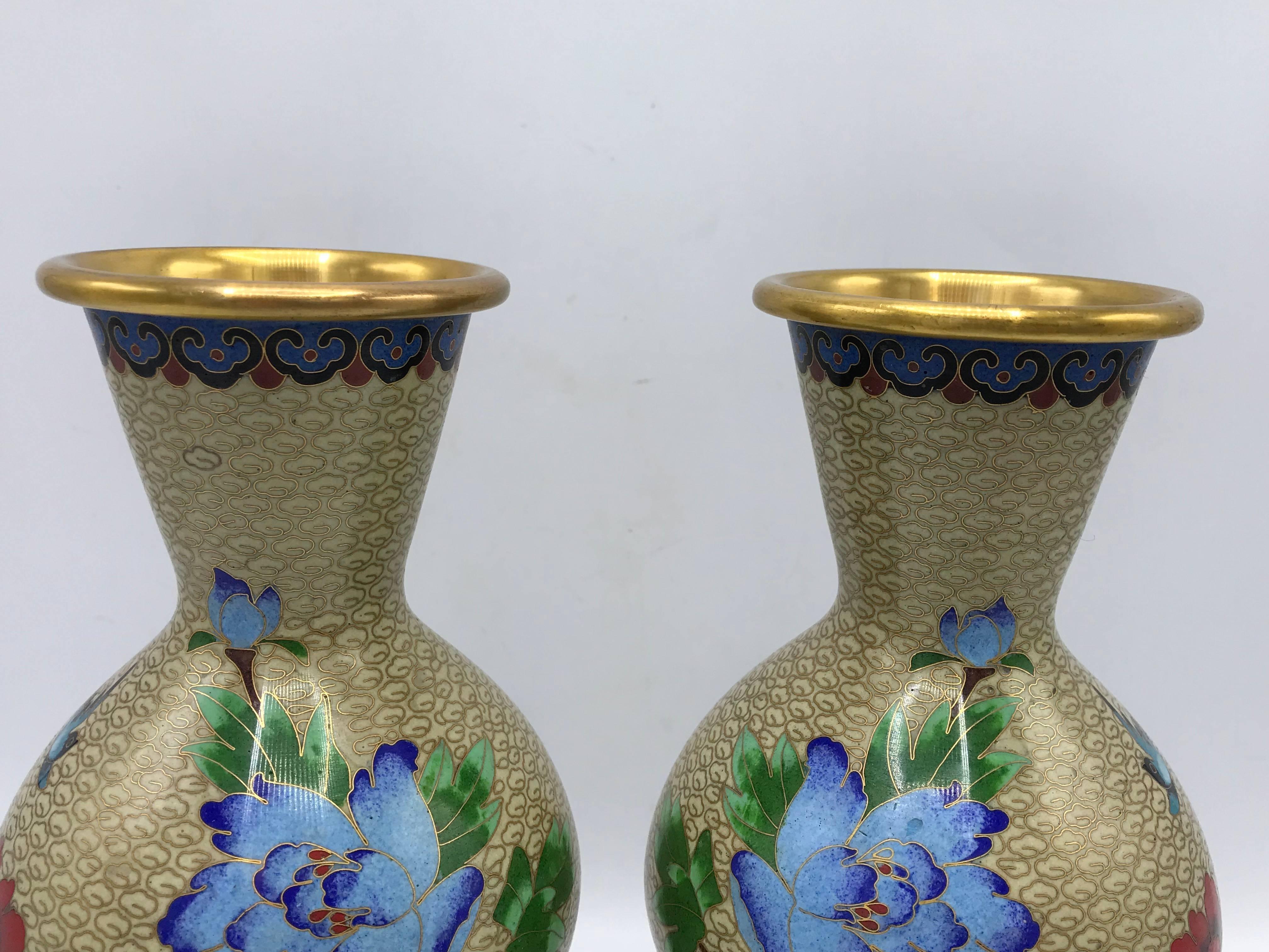 Offered is a beautiful, pair of 1960s cloisonné vases with a polychrome floral motif. Beautiful shades of blues, reds, greens, and purples pop against a soft tan background. The pair includes matching, decorative wood stands. Measures: 8.5