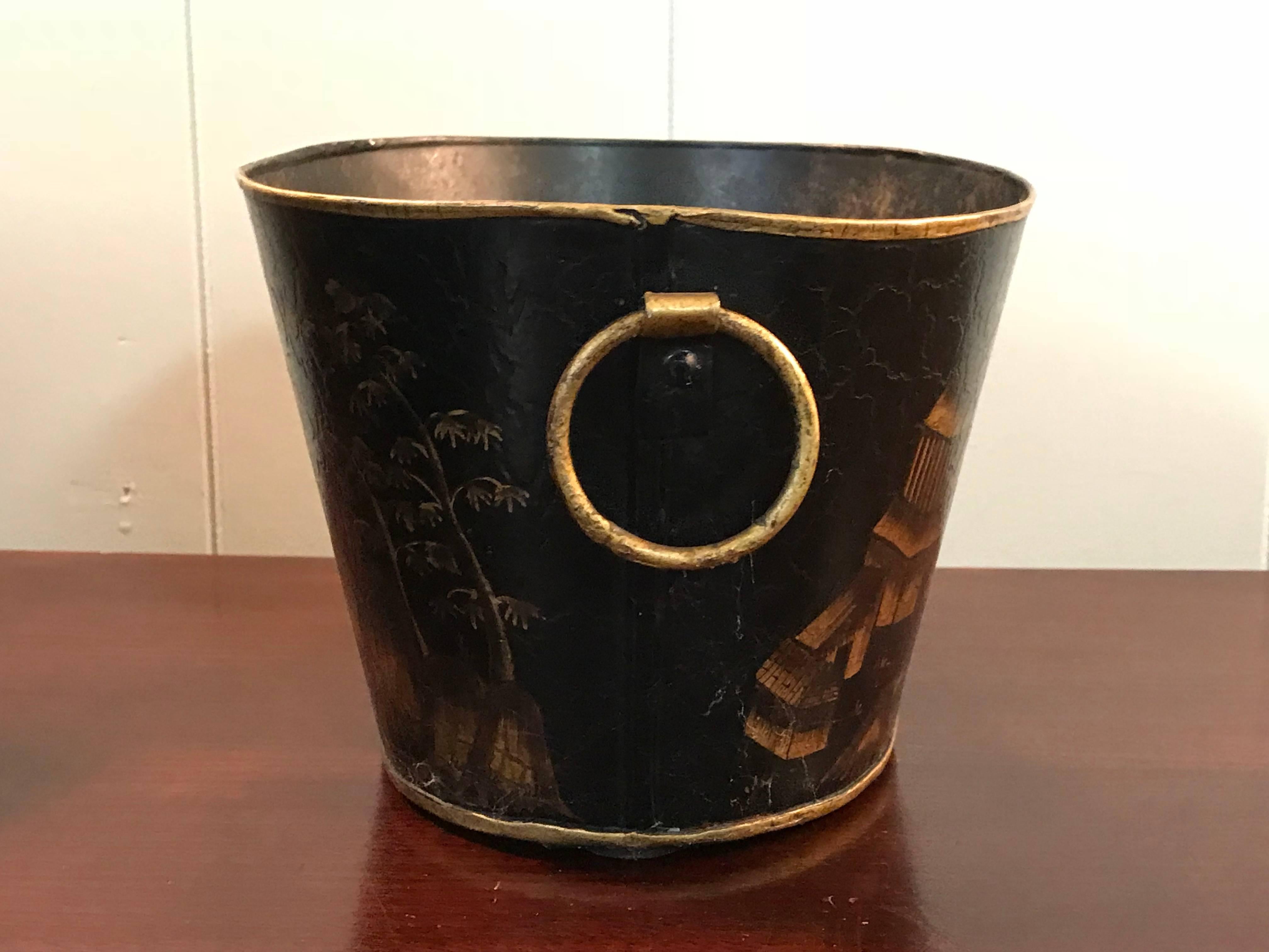 Offered is a fabulous, 1950s black and gold tole cachepot planter. The piece has a stunning, hand-painted chinoiserie motif, featuring mountainsides, pagodas, and ornate foliage. Has two circular, gilt handles - one on each side.