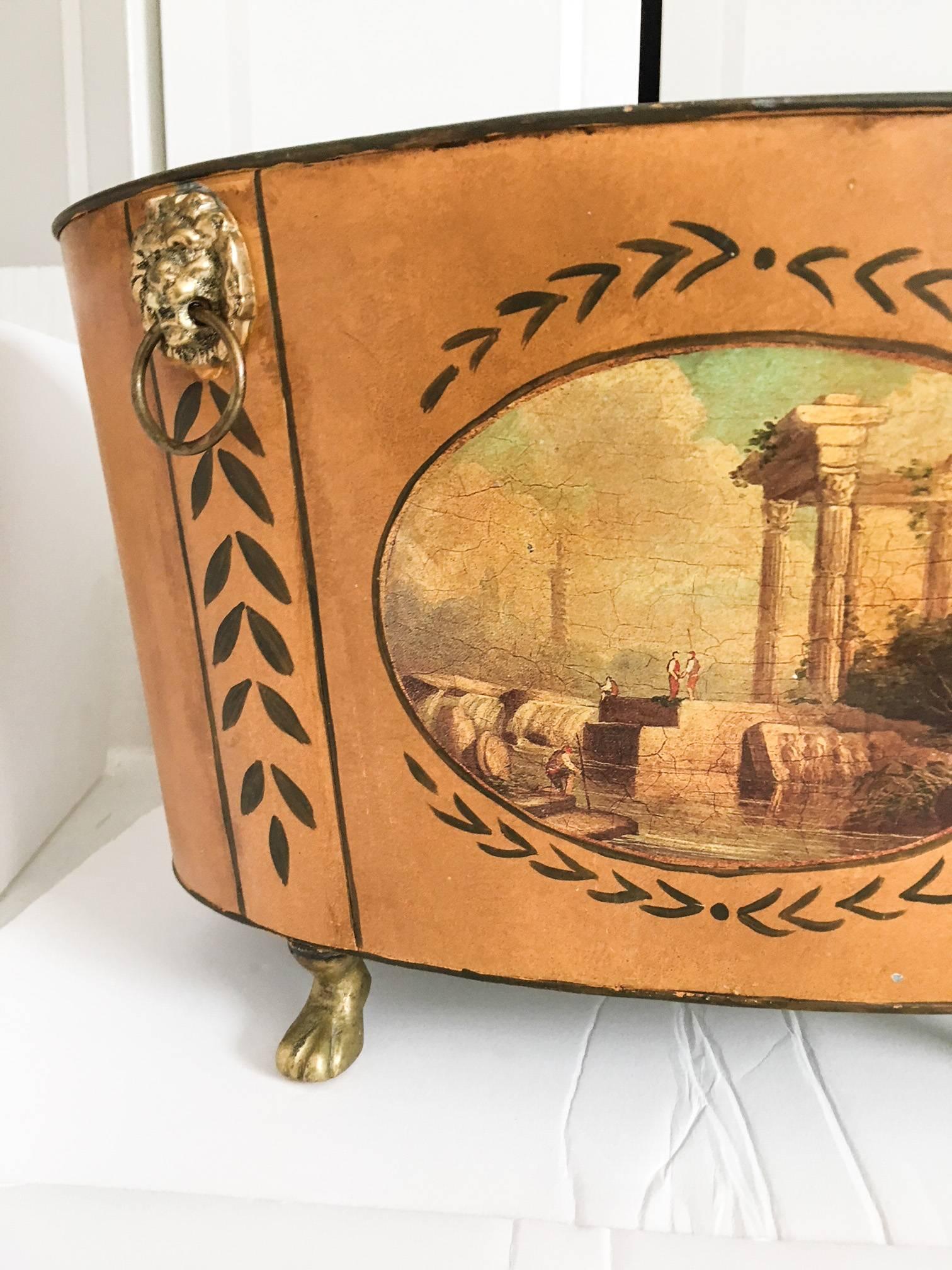 Wonderful butterscotch yellow tole painted metal oval cachepot decorated with brass lions head doorknocker-style handles and hoofed brass feet. Both sides have a decoupage paper Italianate scene; one scene has a small chip at the bottom corner as