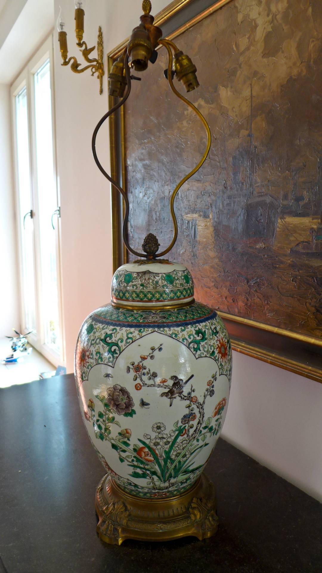A very massive impressively decorated 19th century porcelain Chinese jar converted to a lamp with three bulbs and enamel paintings of birds and flowers and braches with blossoms. The lamp is standing on the inlade bronze base over four legs with