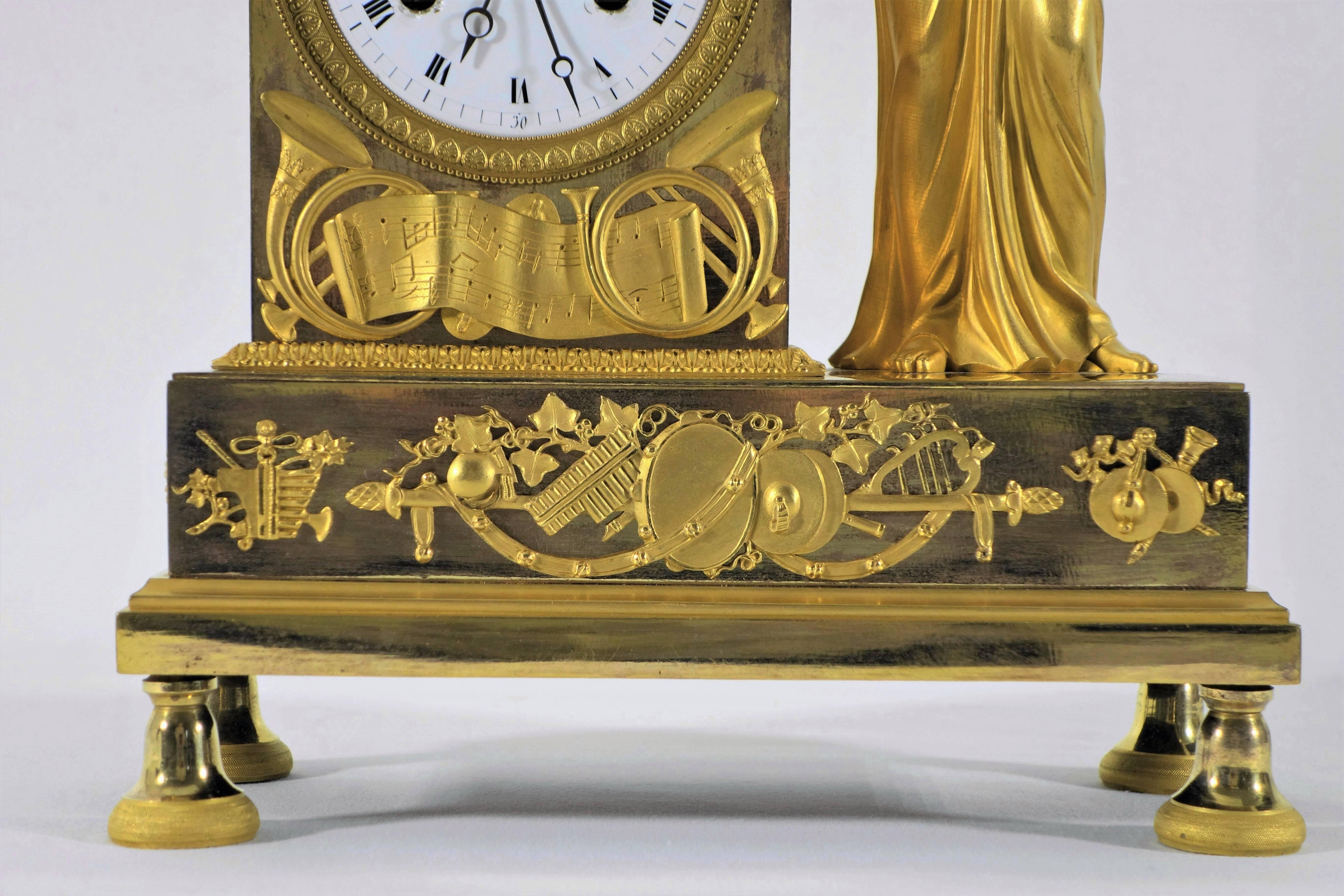 French Empire mantel clock, gilt bronze. A classically dressed female flutist is standing by the clock. The rectangular base is richly decorated with music instruments. Clockwork stamped with AB.
 pendulum and key included.
Dimensions: (H x B x D)