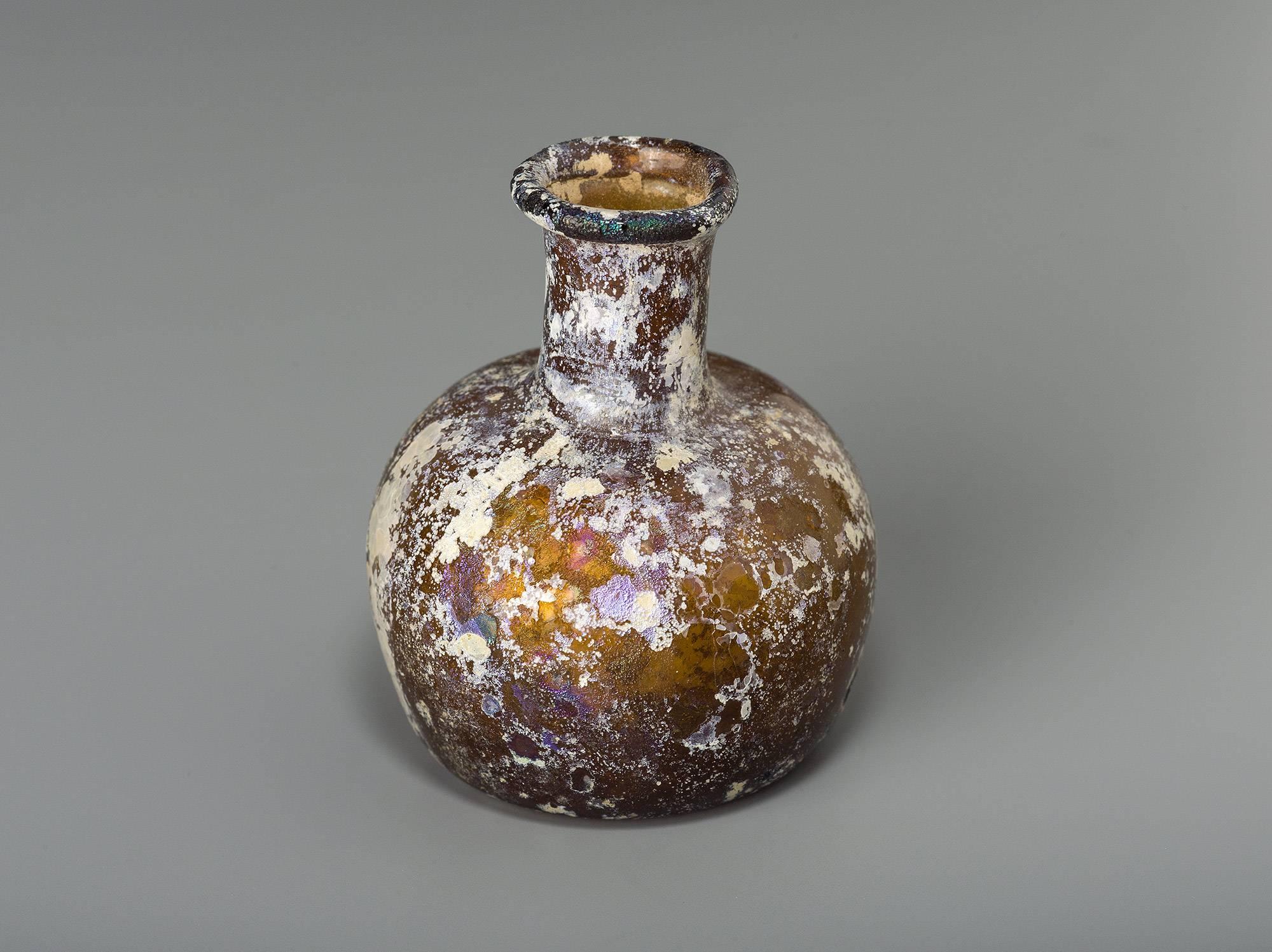 This small size bottle has remains of iridescence all over the body. 
The base is indented and the cylindrical neck is ending with an infolded lip. The bottle is intact and in a good state of preservation.

Provenance:
Private collection