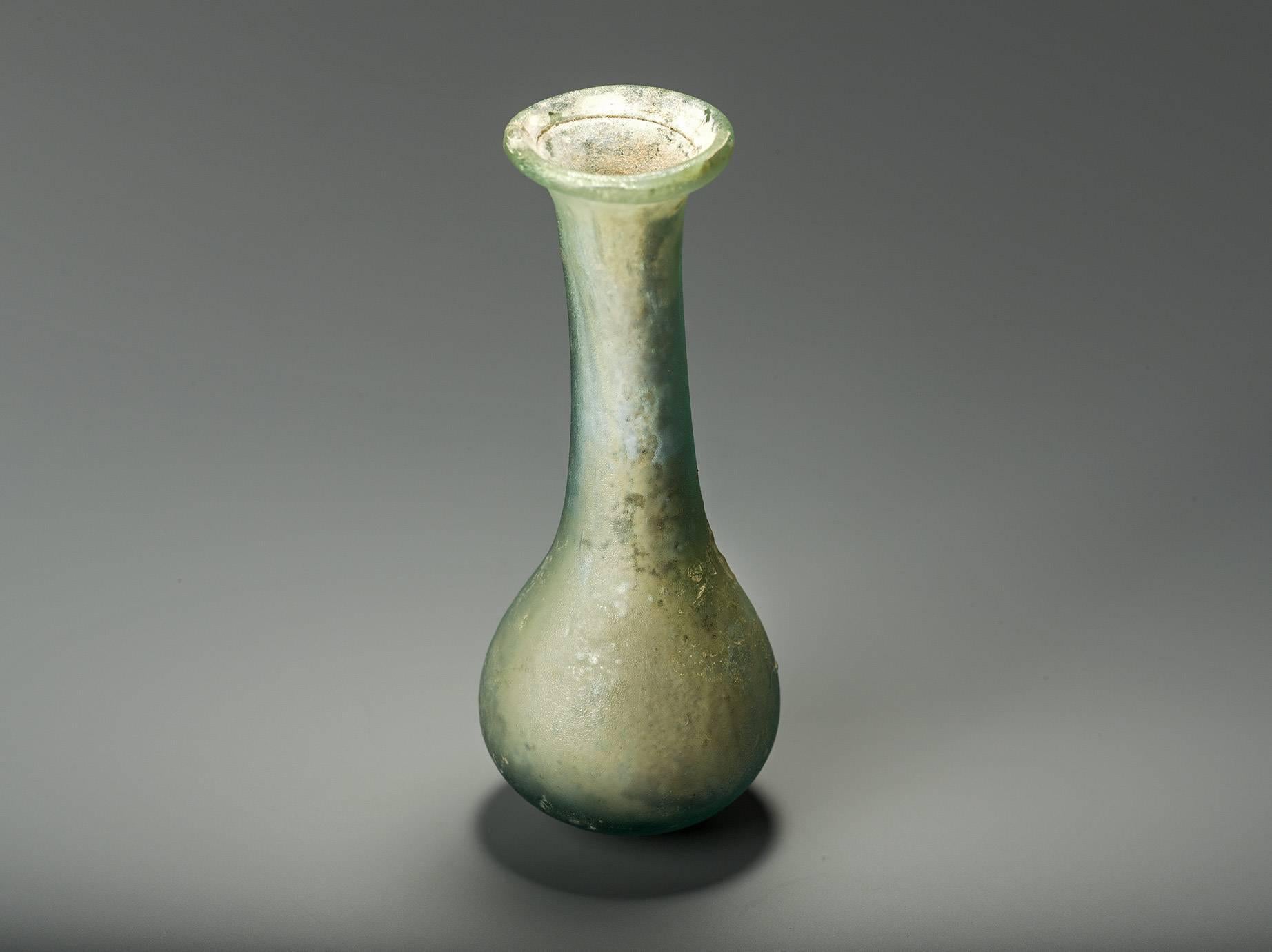 Opaque greyish green glass. Roman, c. 1st Century AD.
This unguent flask has a tall neck that is ending with an inward folded rim. The flask is intact and in very good state of preservation. There is a pin mark, a remains of the glass rod, on the