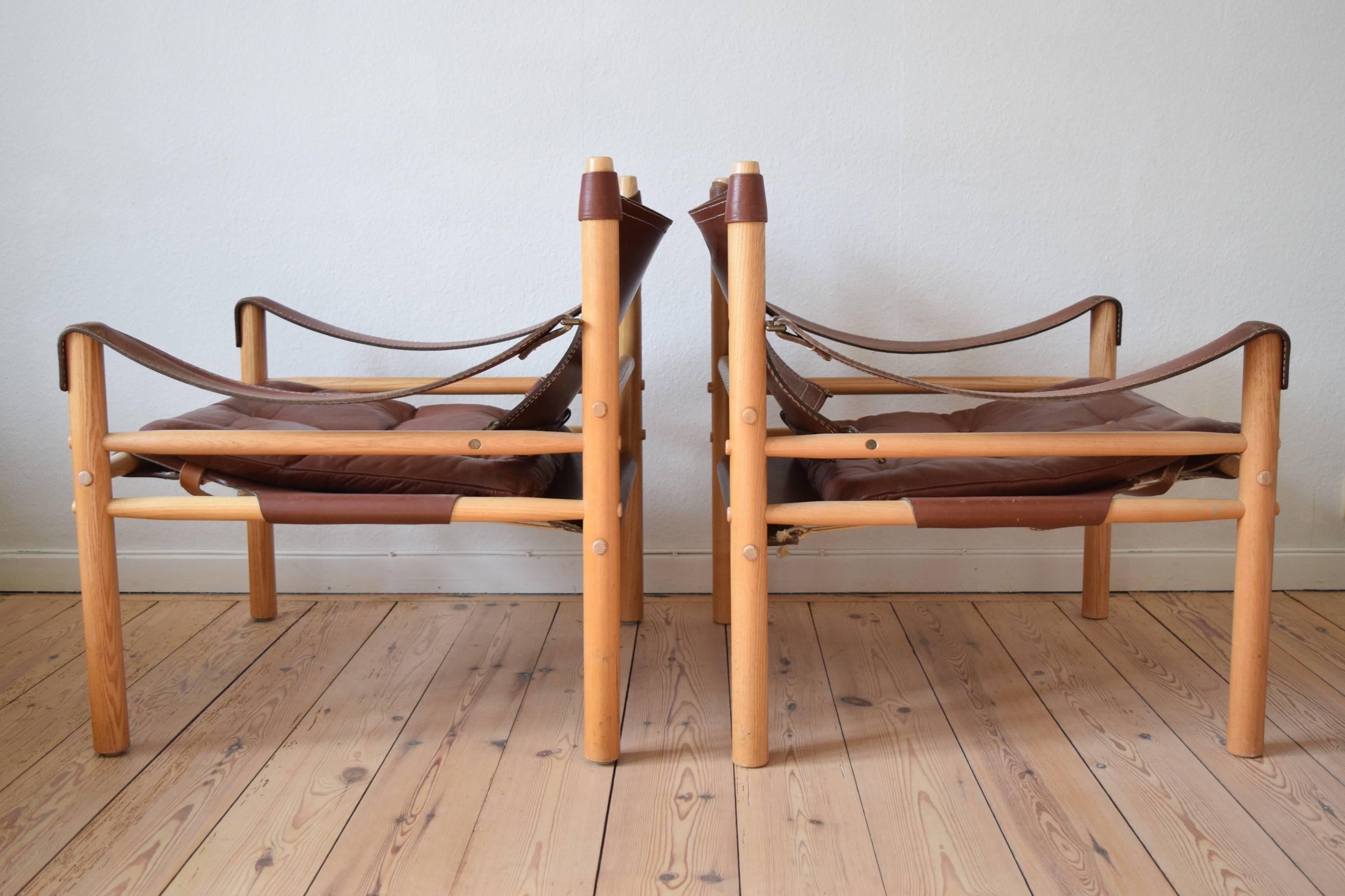 A matching pair of safari chairs designed by Arne Norell and produced during the 1970s by Norell Furniture, Sweden. These chairs feature cognac leather cushions and seats and seats with brass buckles on an ash frame. The seats can be adjusted from