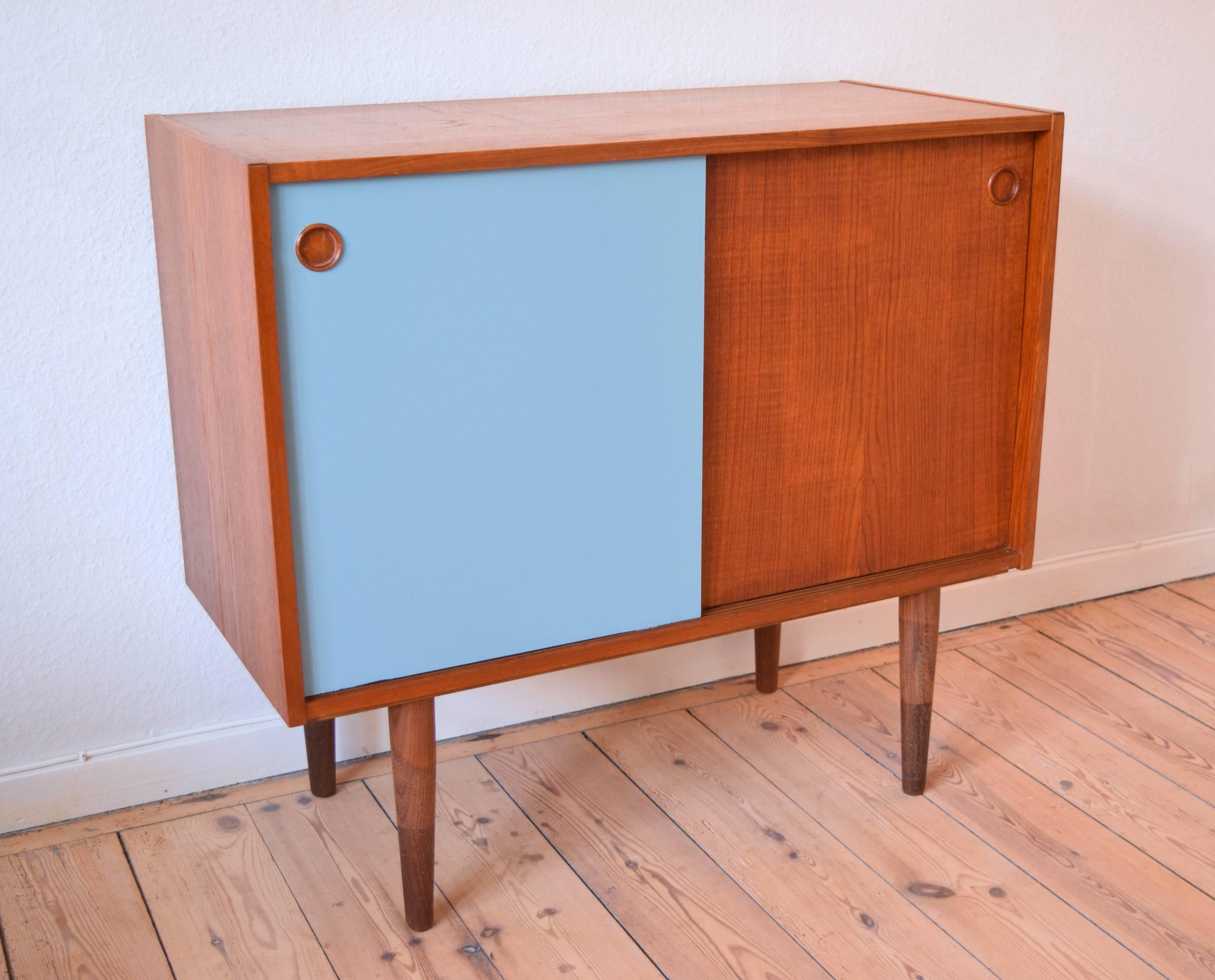 1960s teak sideboard with sliding doors. Features one teak and one painted door. Sits on turned and tapered two-tone solid teak legs. Apart from a few small marks here and there commensurate with age and usage, this piece is in a good condition.