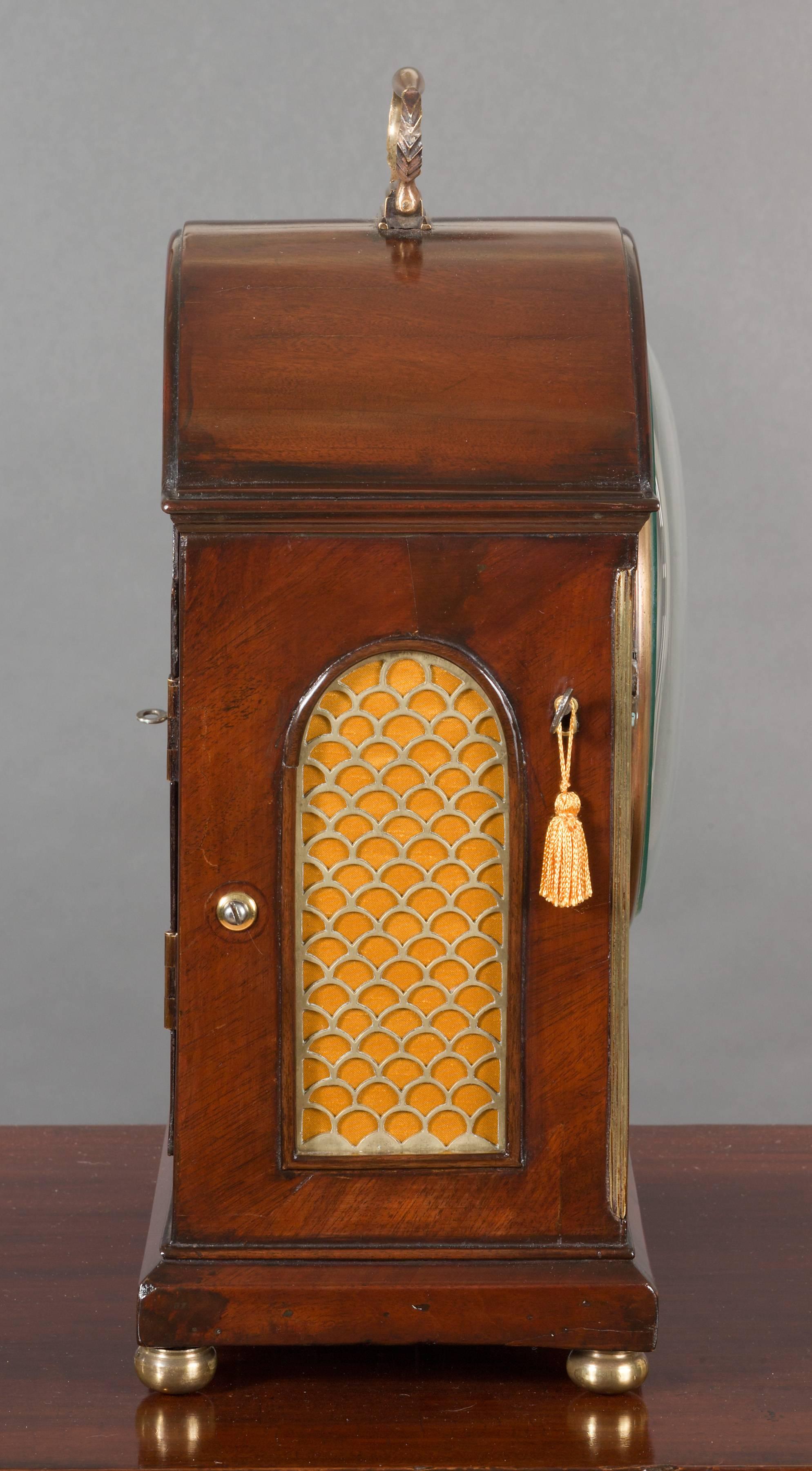 Georgian break arch mahogany bracket clock standing on a raised chamfered plinth and resting on brass bun feet with silk lined fishscale sound frets to the sides. 

Flame mahogany front inlaid with brass decoration, surmounted by a brass carrying