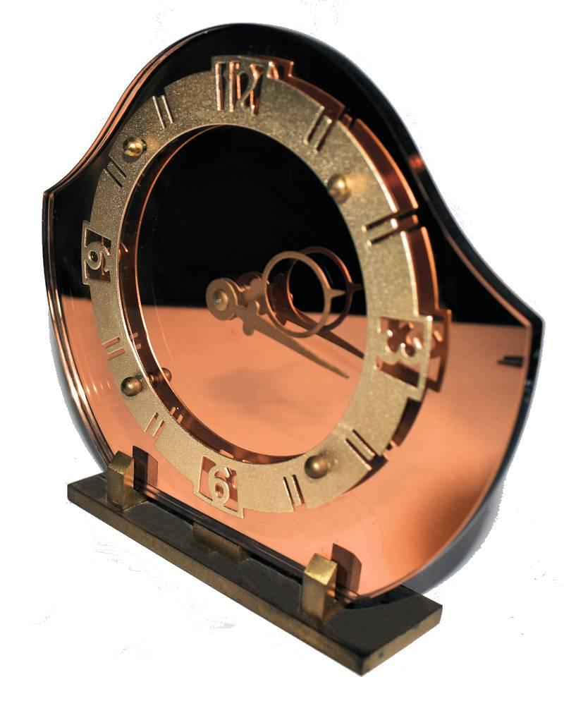 Wonderful 1930s Art Deco English mantel clock. This clock is an ideal size for mantel areas or desks. Features a thick polished edged mirror clock in a peach color with a gilt metal stylized frame and numerals, all of which sits on a solid brass