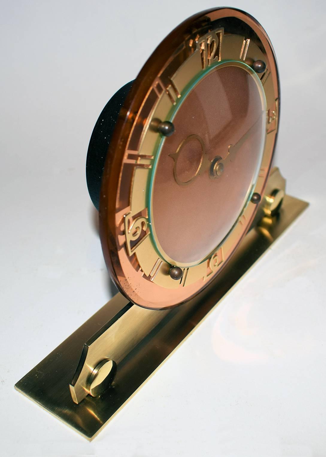This is a very attractive English Art Deco clock, made circa 1930. It has a eight day wind up movement. The base is brass, the bezel is also brass with gold tone finish. The backing glass is peach-coloured mirror . In excellent condition. Full