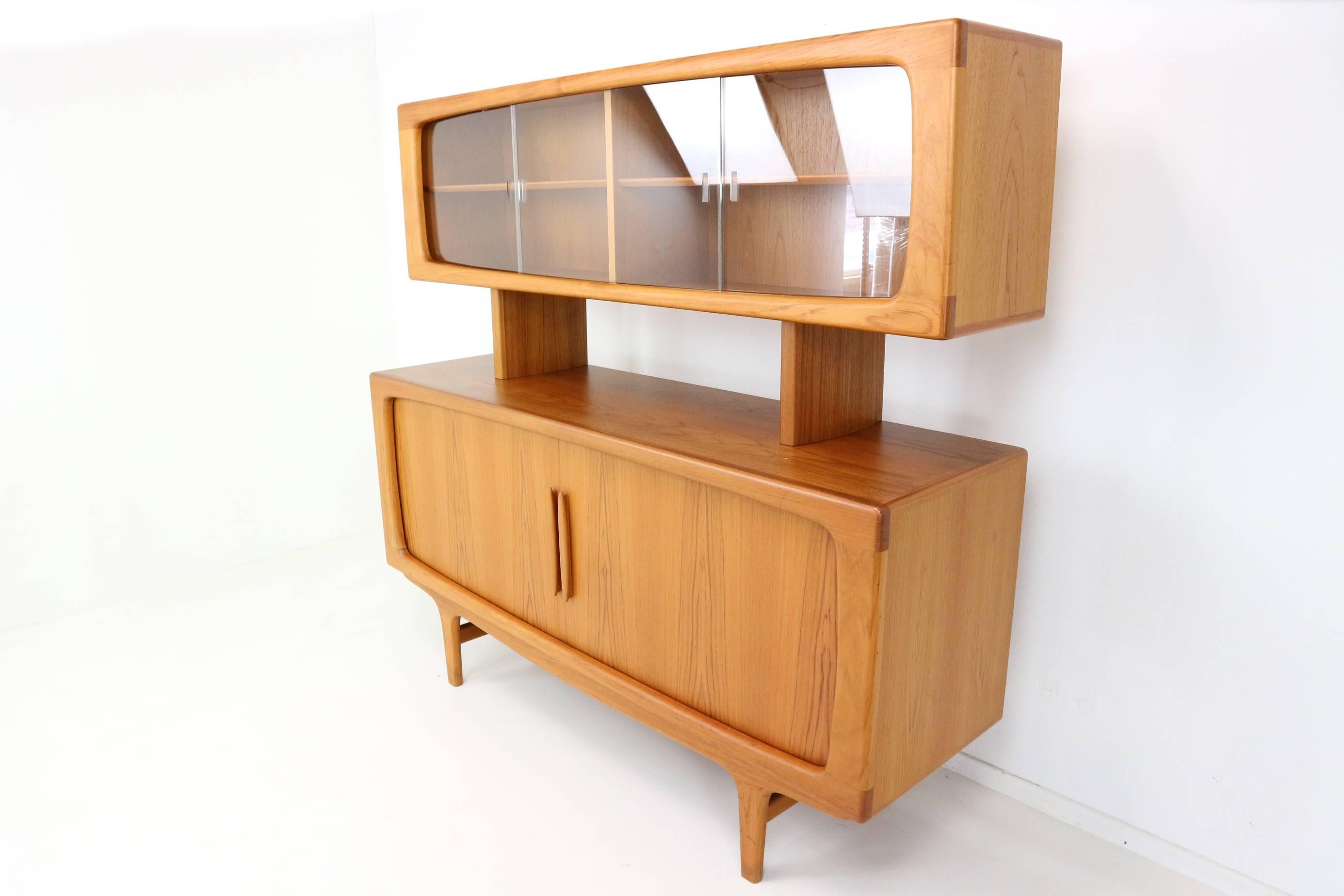 Designed by Johannes Andersen and constructed in Denmark by CFC Silkeborg. This unadorned piece exudes both quiet beauty and functionality. The teak doors are tambour and open to reveal shelving and drawers for storage. The top part has glass