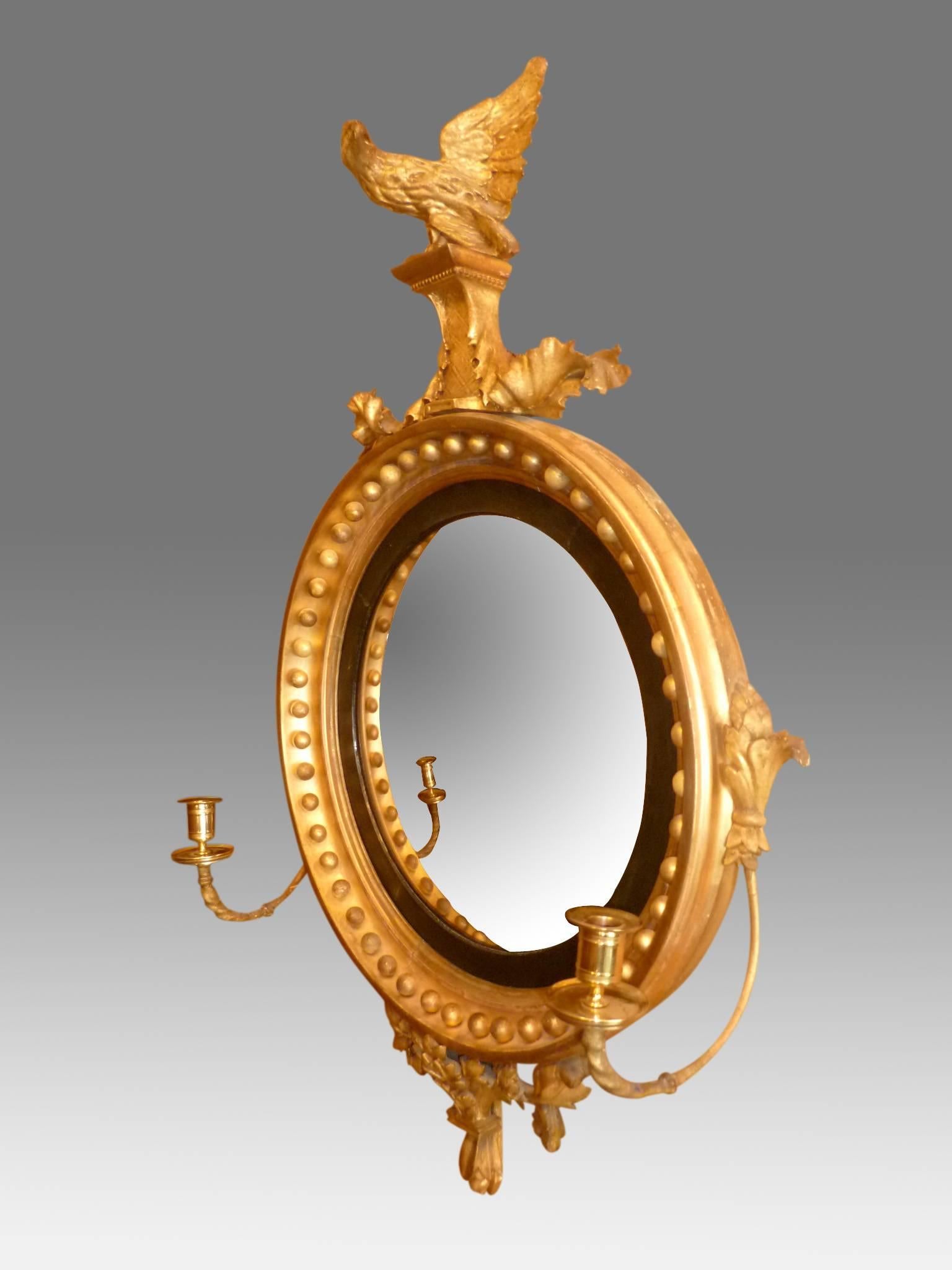 A fine Regency period giltwood convex mirror with carved eagle surmount, perched on a raised platform flanked by carved oak leaves. The reeded ebony sight edge surrounded by gilt balls, with tied leaf pelmet below. Fitted with original scrolled arms