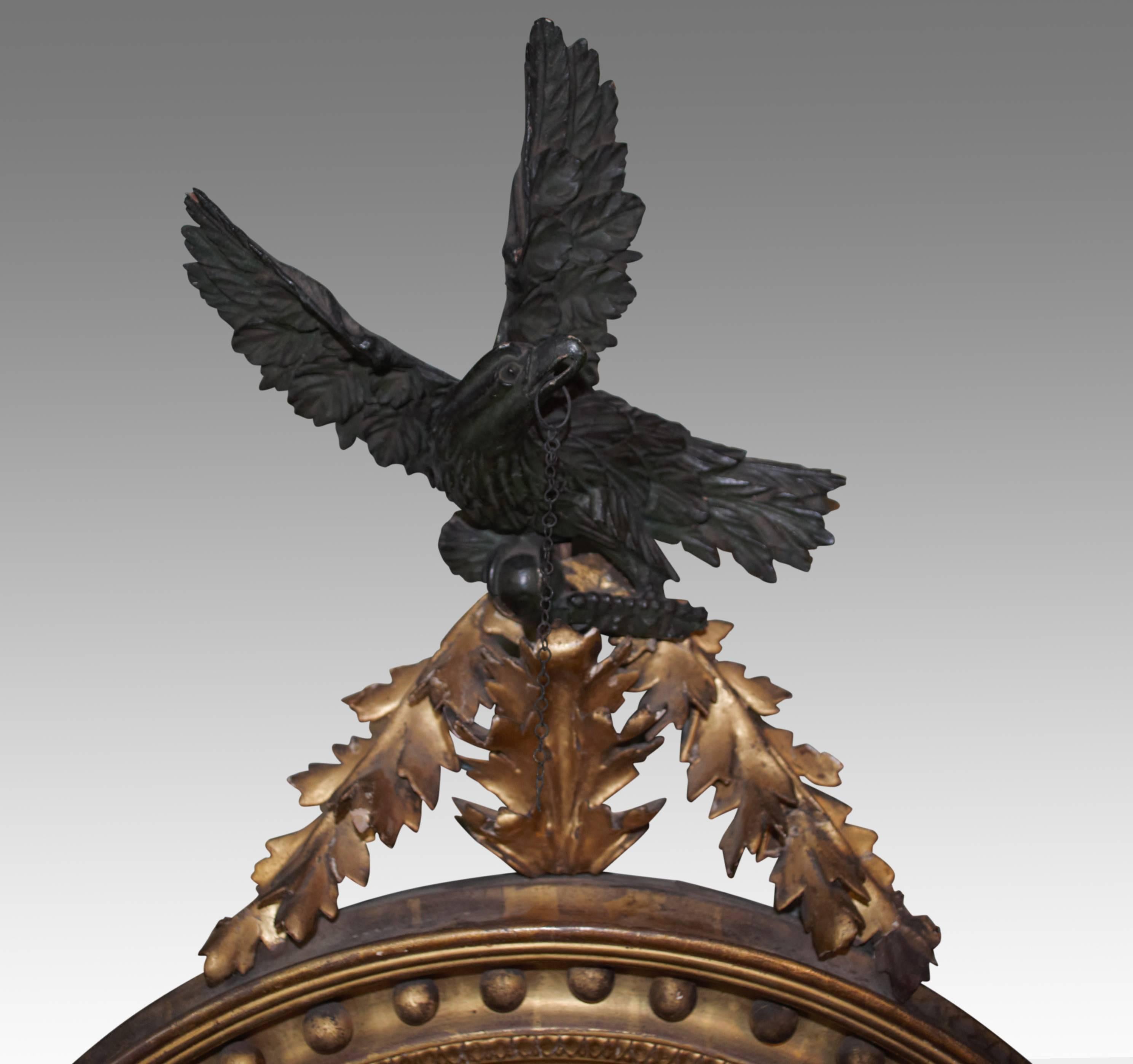 A fine Regency period giltwood convex mirror with carved ebonised eagle surmount, perched on a raised leafy pediment. The scooped frame set with gilt balls, with half sunburst pelmet below.