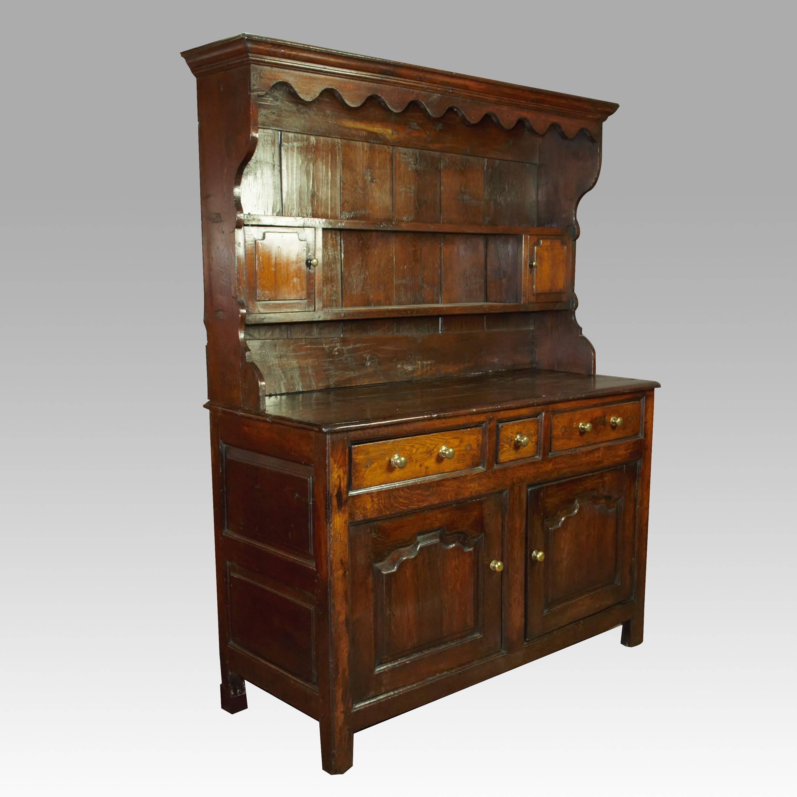 A fine quality Carmarthenshire, 18th century oak dresser and rack of small size and excellent color. The wavy-edge canopy above plate racks and two small cupboards, raised on a panelled two-door cupboard base with three drawers and standing on stile