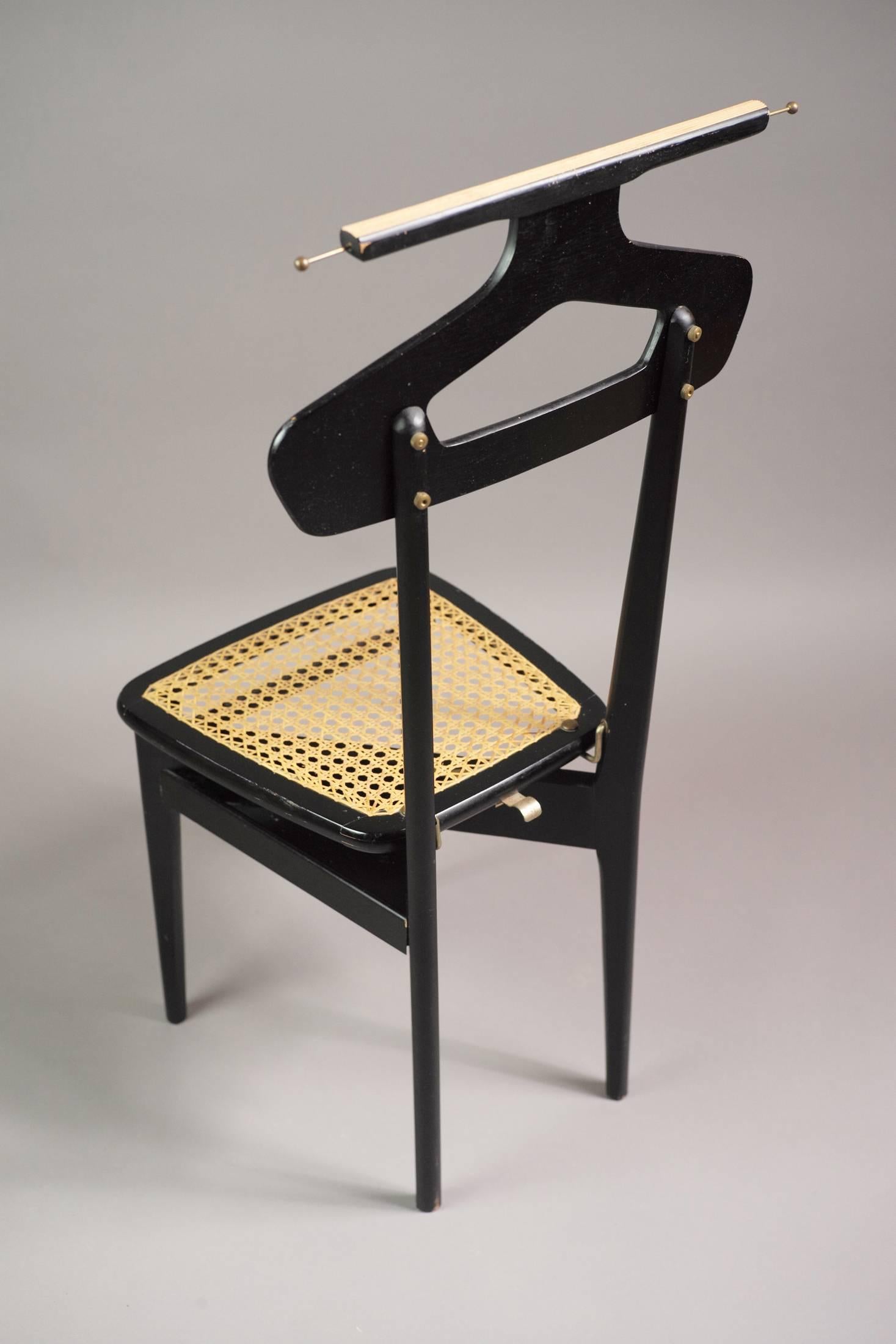 This valet was produced by the Italian design house Fratelli Reguitti in Italy, circa 1950. It is made from ebonized wood with a removable cane seat. It has four brass nuts and bolts, two retractable tie hooks and a spring that allows the raised