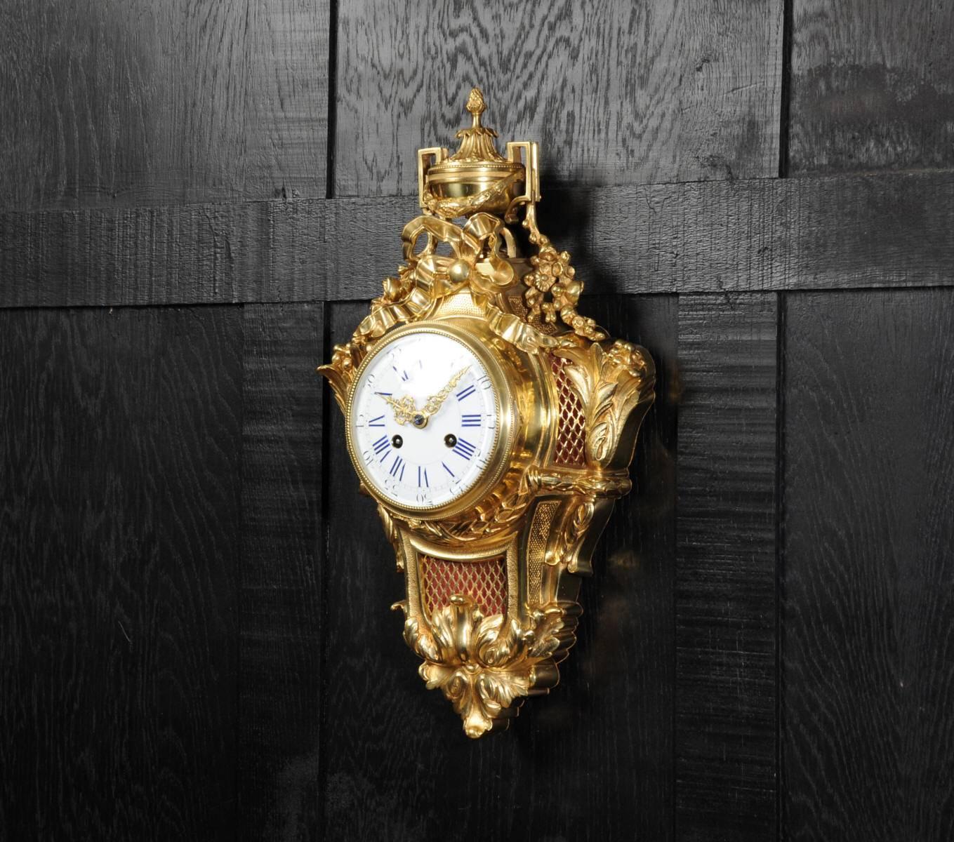 A very elegant Cartel clock, Louis XVI in design with a handled urn and acanthus decoration. A large bow sits above the dial and sound panels to the sides and front allow the delicate sound of the bell strike to be heard. It is finely made in gilded