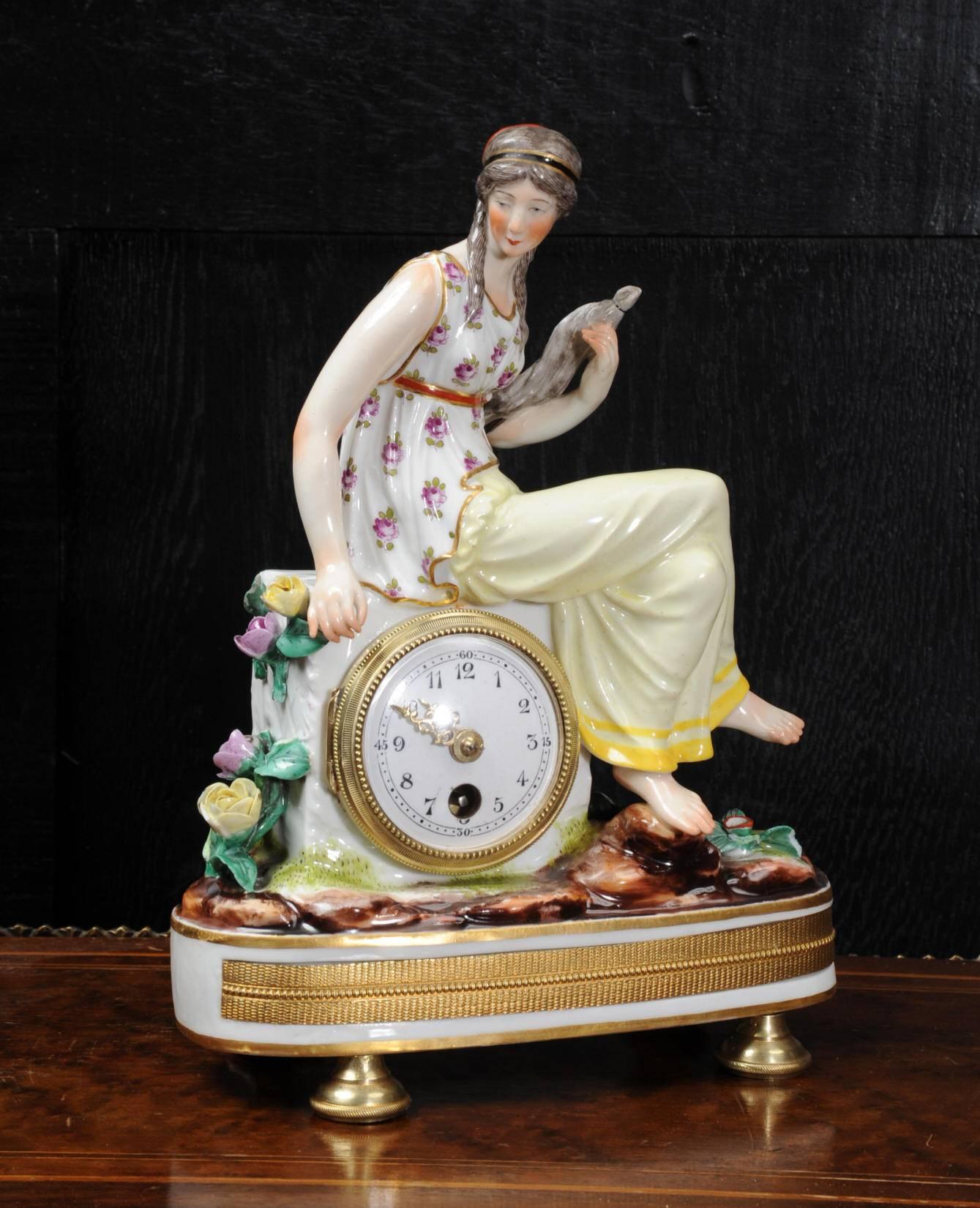 A beautiful Royal Vienna type porcelain boudoir clock featuring a classical maiden seated on a rock. She is holding a dove and is surrounded by delicate flowers. The tiny French movement is mounted in the rock and has an enamel on copper dial with