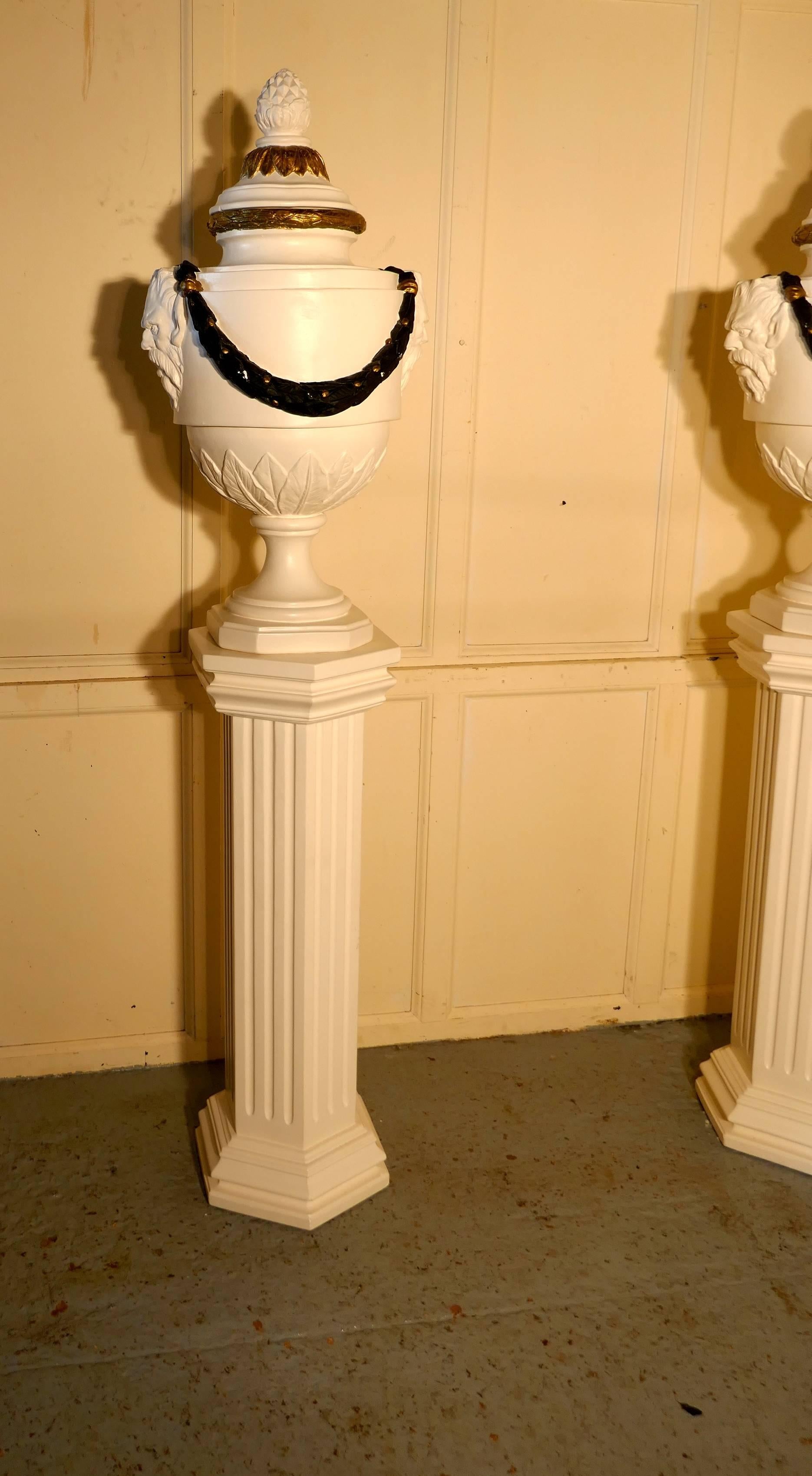 A pair of French Urns set on Classical column pedestals 

This is an imposing pair of Classical tall lidded urns, they have the mask of a greek deity on either side adorned with a laurel wreath, the painted decoration is in black and gold
The