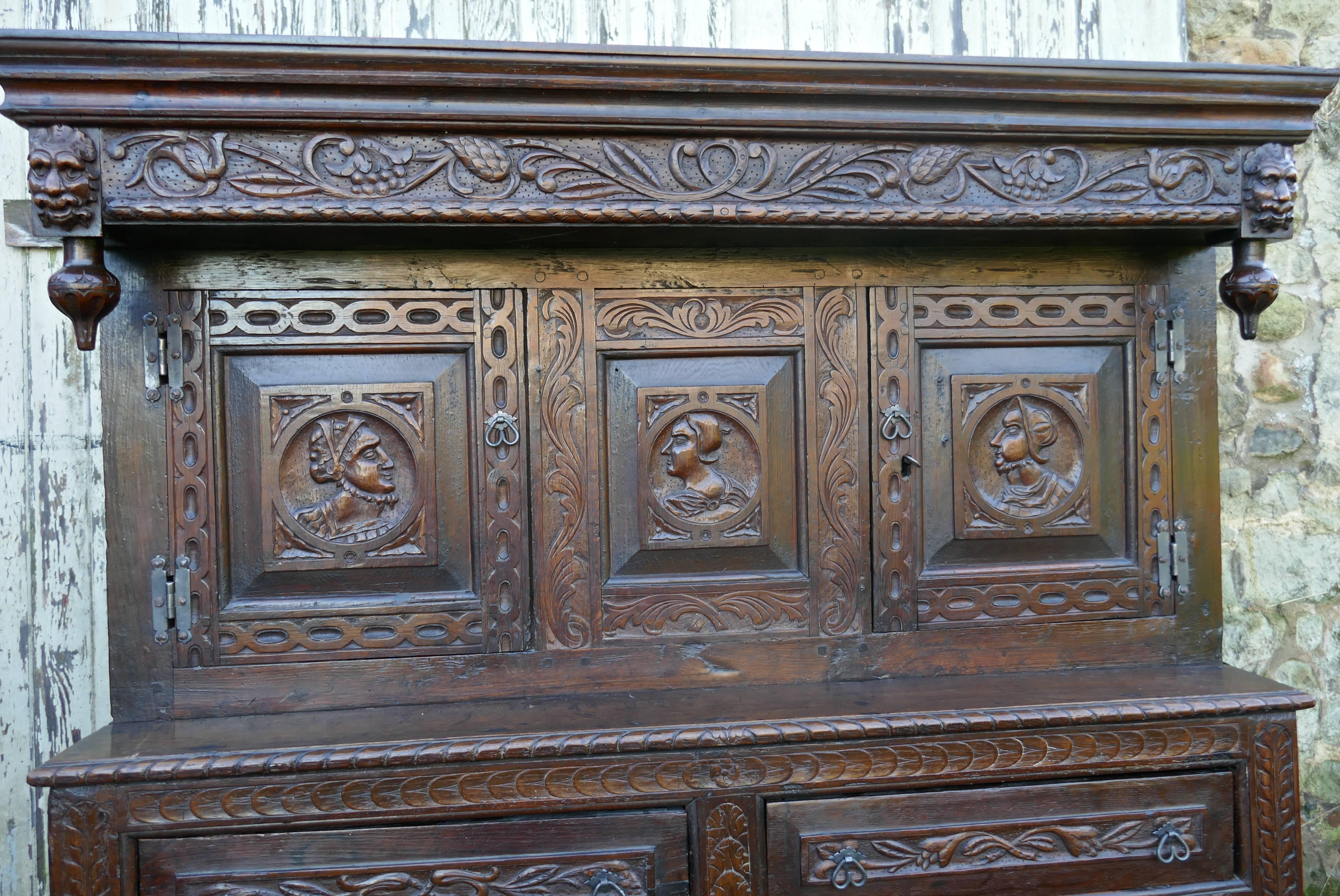 This is an old and charming rustic piece of carved oak furniture, the top section has a long cupboard enclosed by 2 doors and a centre panel, each of the carved panels has the face of a warrior or similar character, it has a carved cornice on the