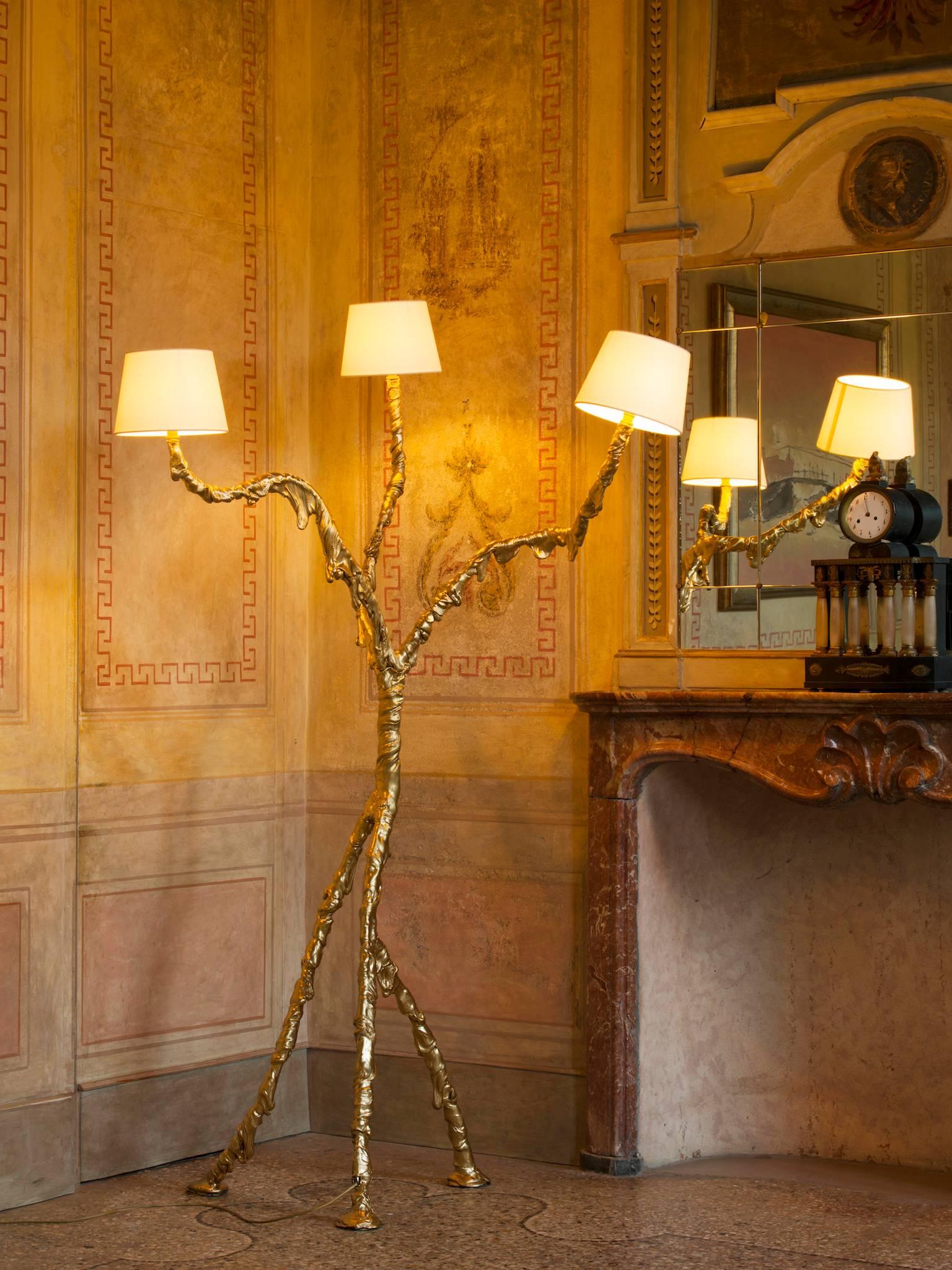 It is a polymorphic lamp. Sinuous and slender with very long legs. It is precious, coated with “pure gold.” It has a stem that branches out like a tree, the dynamics and proportions of an inverted chandelier. The light is diffused by three small