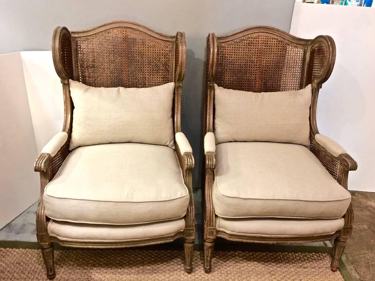 This is a rare pair of early 20th century double caned Louis XVI-style wing chairs that retain their original grey-green paint and which have acquired just the right degree of patina, The cane is in good condition without any noticeable damage.