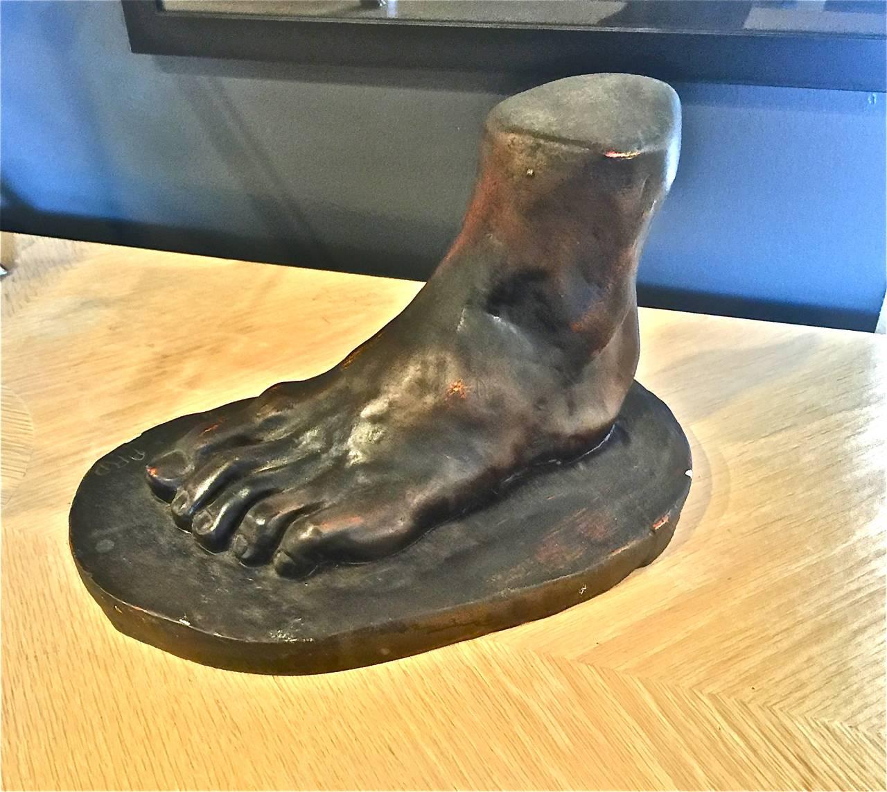 These are early 20th century French plaster castings of male feet in the neoclassical style. One of the feet features a bronzed patina and the second foot is in patinated paint. One of the pieces is signed with initials. Both casting are in overall