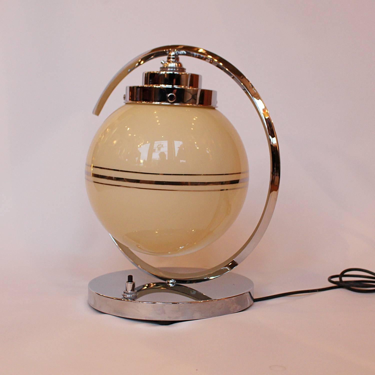 An Art Deco table lamp. Original glass shade with silvered banding, set in a c-shaped, chromed metal frame with stepped top.

Fully refurbished, re-wired and re-chromed, with some replacement parts.

Dimensions: H 34cm, W of shade 20cm, W of base