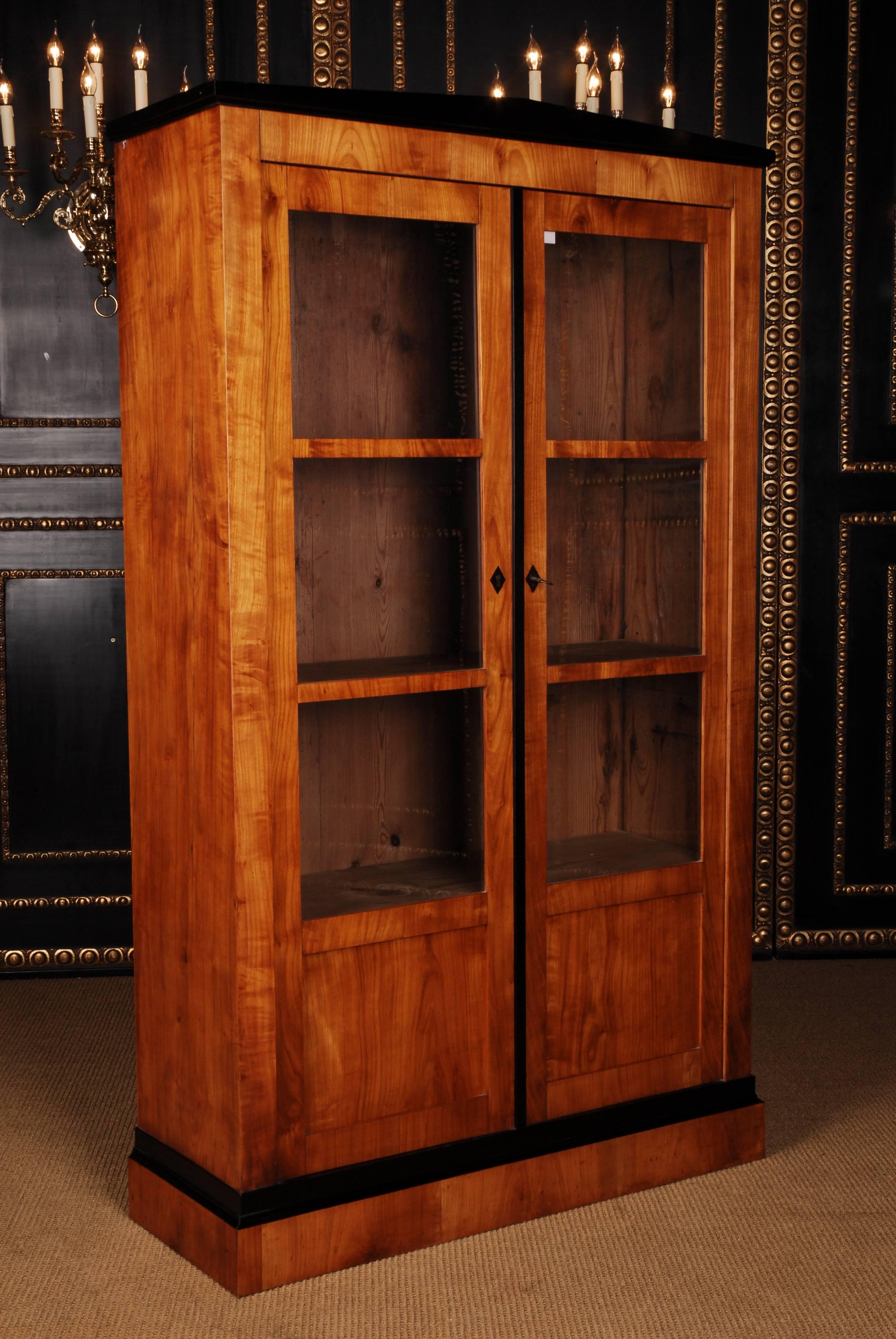 Biedermeier bookcase or vitrine, circa 1815.
Cherry tree on solid partly ebonized. On stepped frame base, high-rectangular corpus, partly blackened. In the front two triple-sectioned, sprouted-glazed, casted doors. Over cantilevered profile cornice