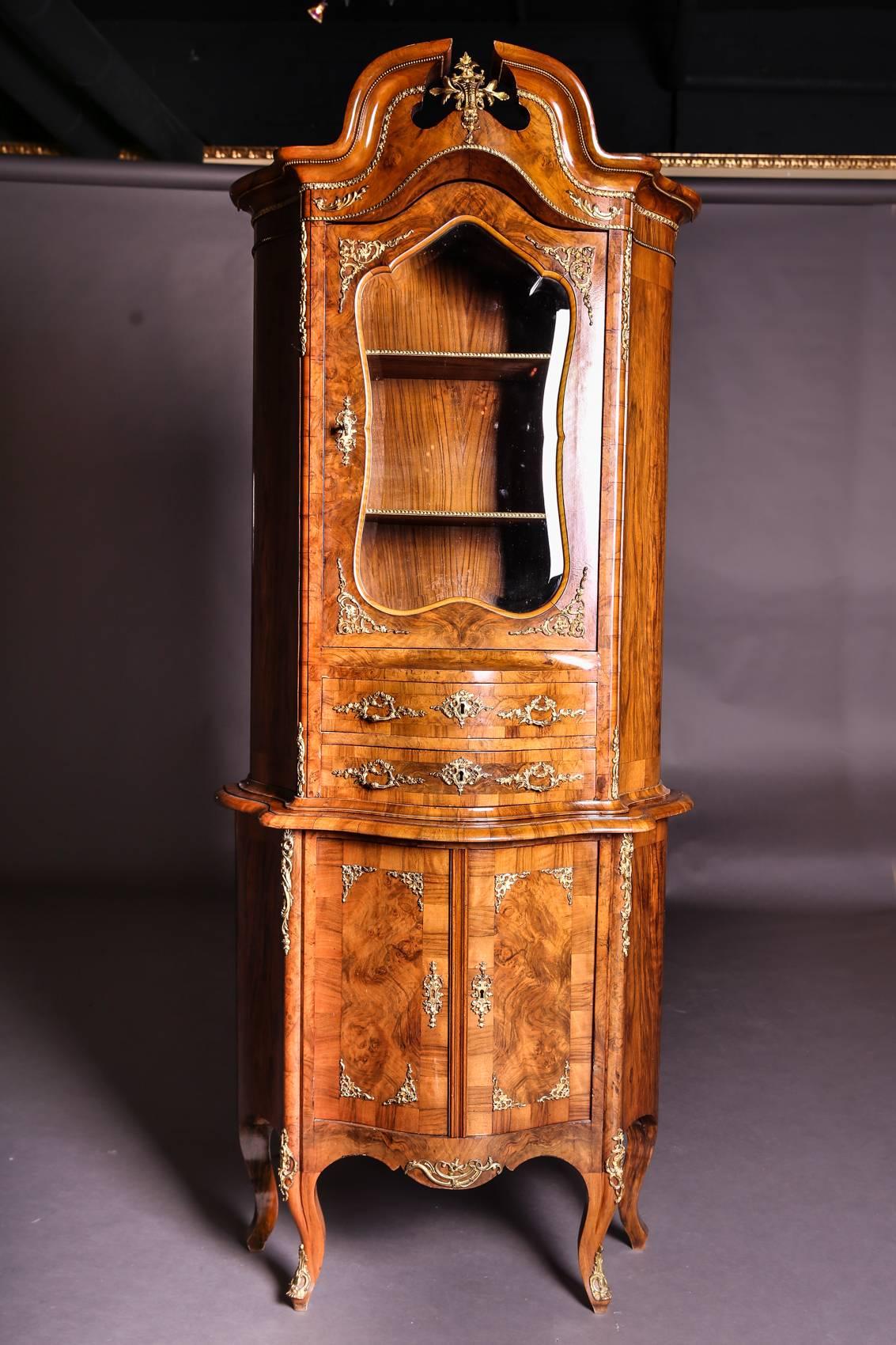 Vitrine in Dresden Baroque. Walnut freed and felted. All-round slightly curved body with rich, gilded bronze motifs. Prefabricated, cracked gable with bronze bead contour. Cut-out glass door over two drawers. Two-door base on high curved