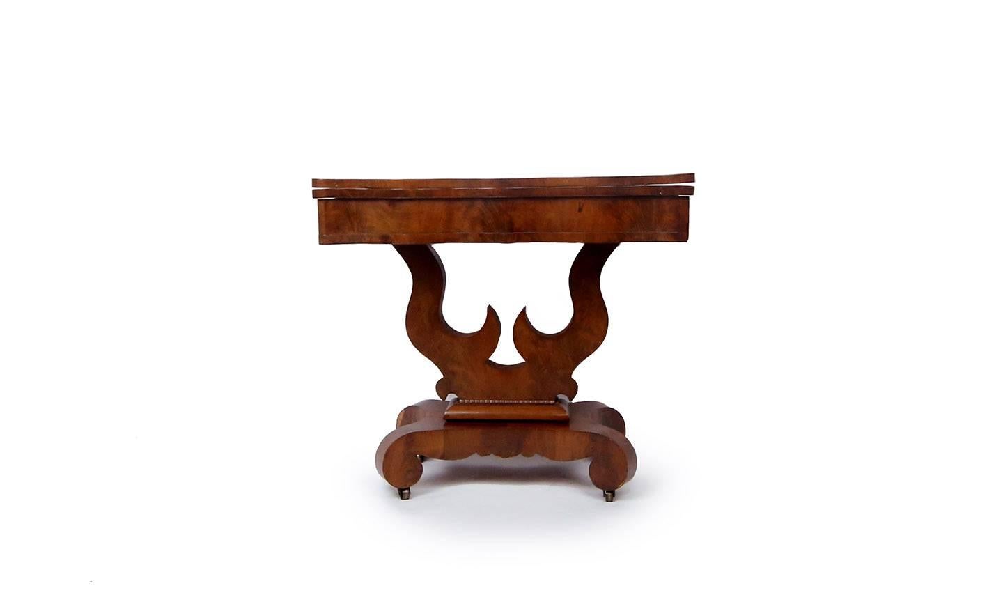 Handsome Empire card or tea table with centre pedestal in mahogany and matchbook flame mahogany. Hinged top opens to reveal compartment and pivots to become square table. Scalloped edge top and frieze supported by bold harp base and scroll legs with