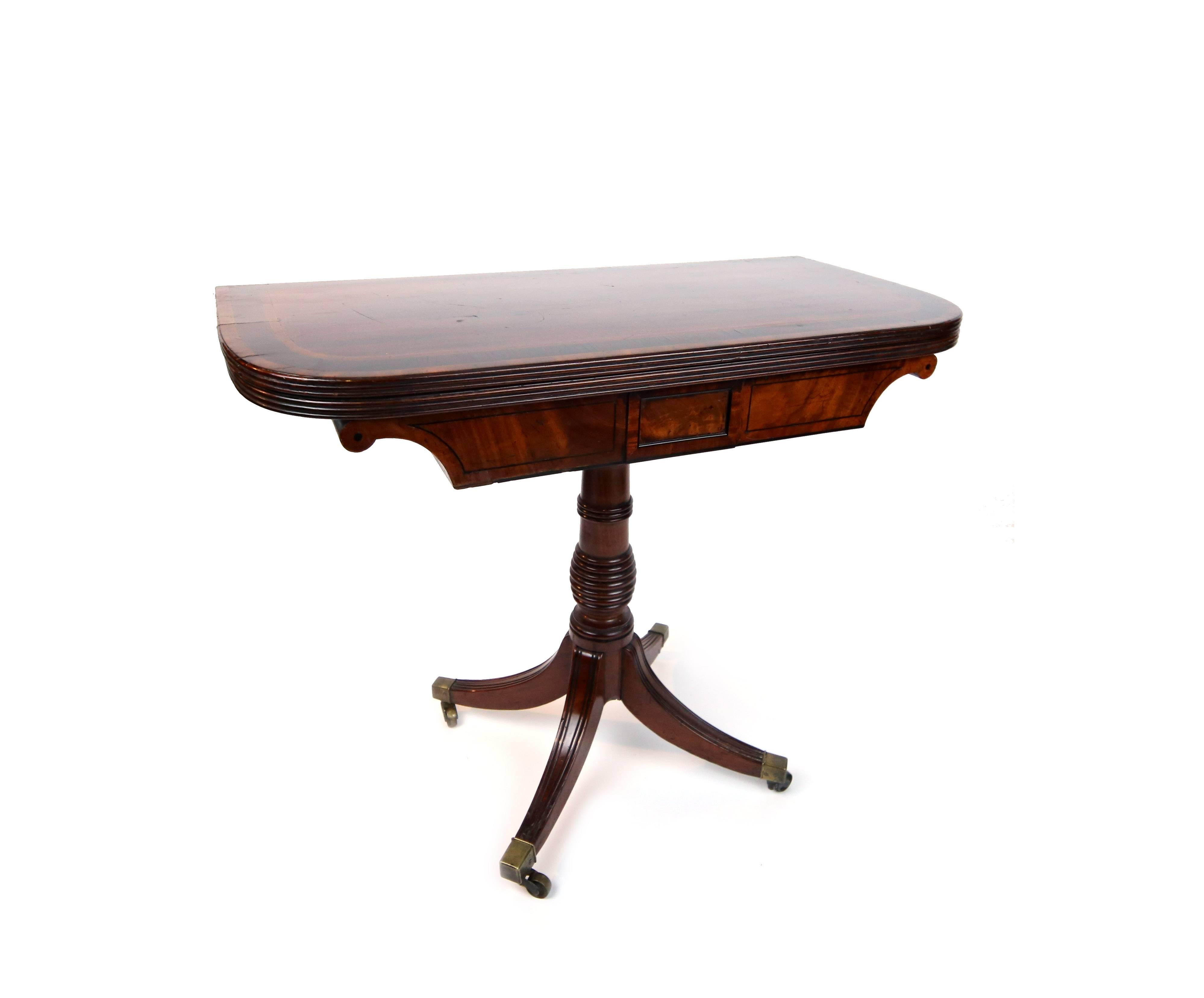 Fine flip-top game table in mahogany, satinwood, and ebony stringing. Cockbeaded table edge opens to original green felt gaming surface. Simple beehive turned pedestal base supported by four saber molded legs terminating in square cap brass castors,
