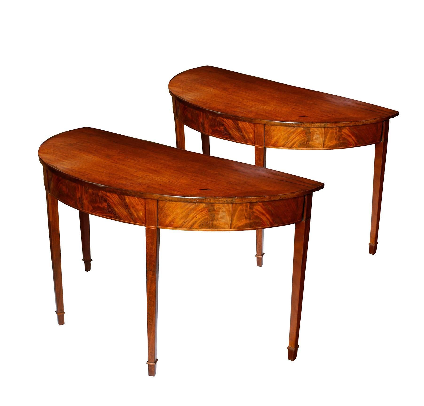 A lovely pair of American console tables in honey colored mahogany.  The two demilunes feature a graceful elliptical shape with flame mahogany veneer on the frieze and cross banding of holly and mahogany.  The tabletops are composed of single