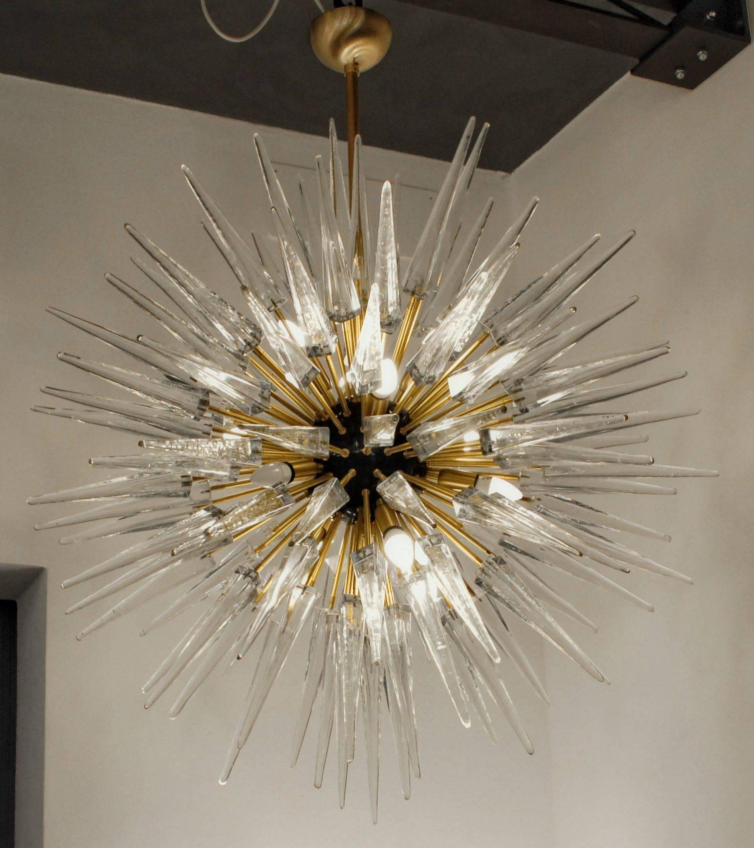 Imposing Sputnik chandelier with spike prismatic shaped glass elements hold by a brushed brass rod. Murano glass pyramid in clear. Made at Alberto Dona’s factory.
Glass is shaped first on the bronzino and then pressed from the punty rod down in a