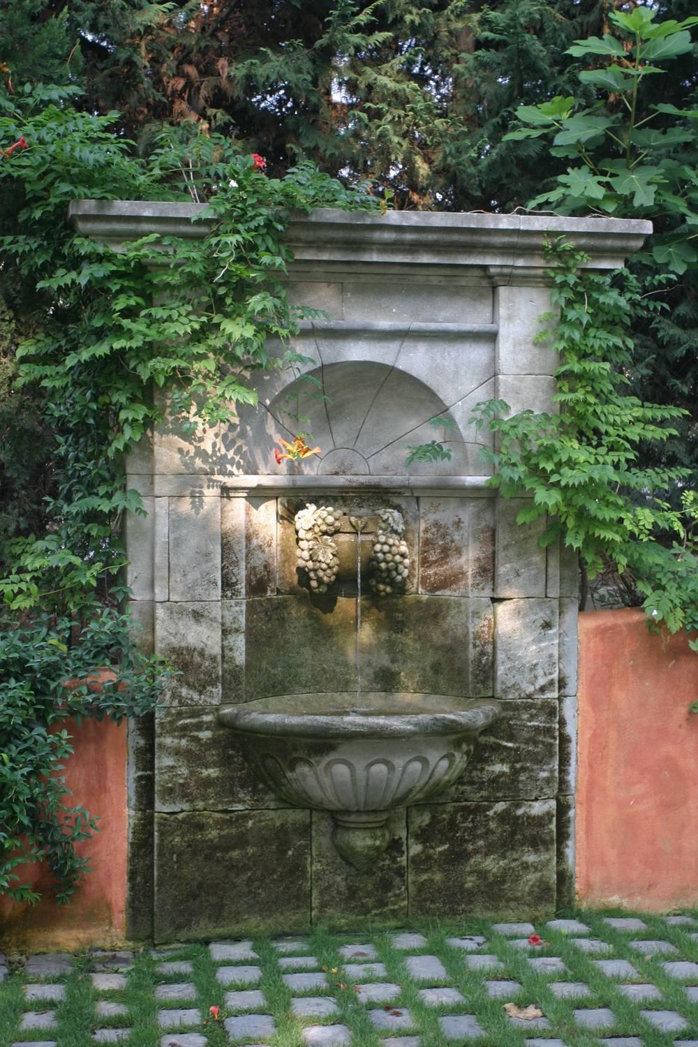 This beautiful garden wall fountain, ideal to lean against a wall, is a piece of fine work-of-art. It shows a large bowl decorated with gadroons and, as a water outlet, a gargoyle finely sculpted by hand with bunches of grapes and vine leaves in a