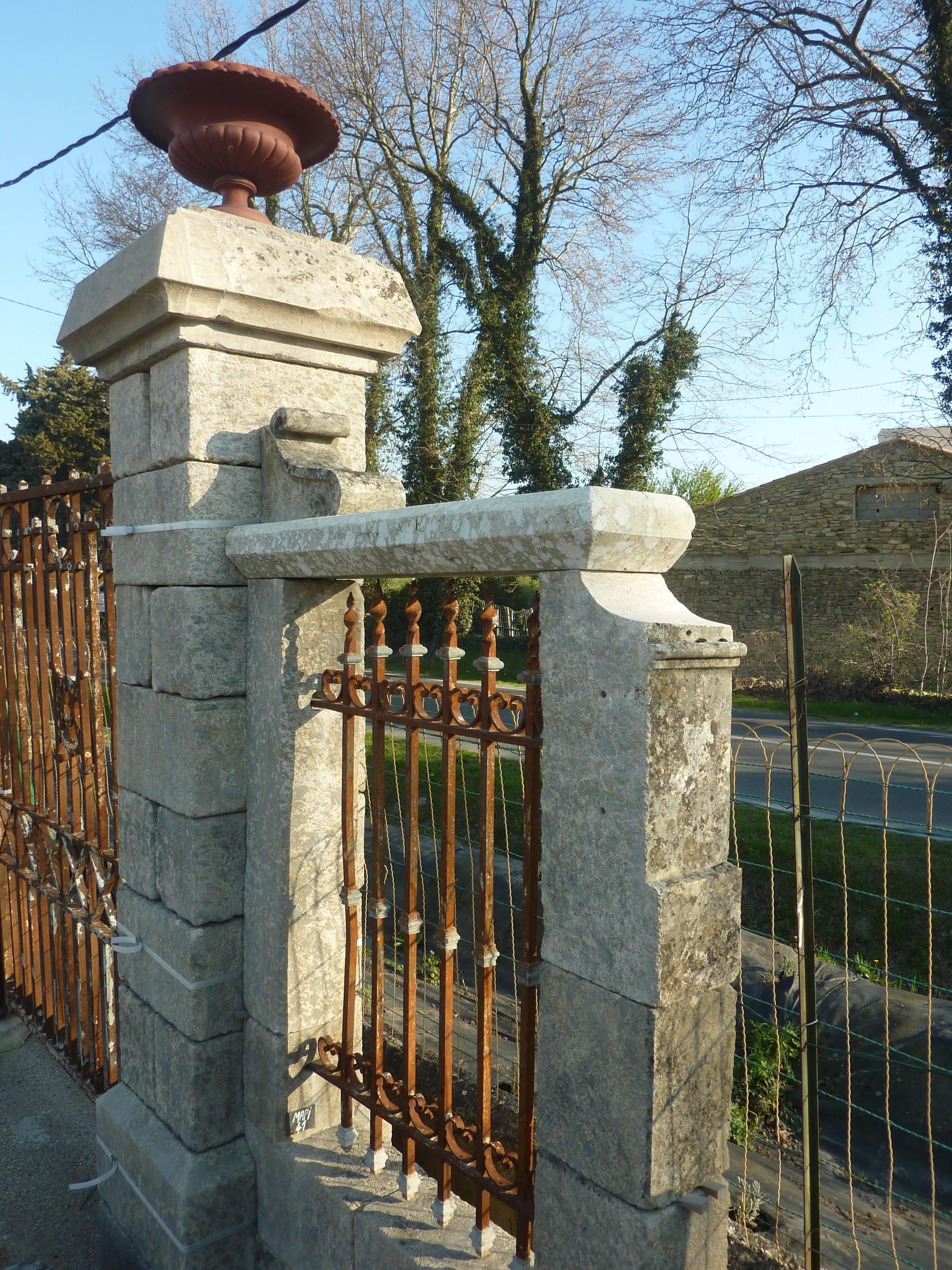 This pair of gate pillars in stone is flanked by long and thin stone low-walls. Each of these elegant pillars is topped by a lovely antique cast iron Medicis vase. This set features two side wrought iron grids animated with volutes and twists.
The