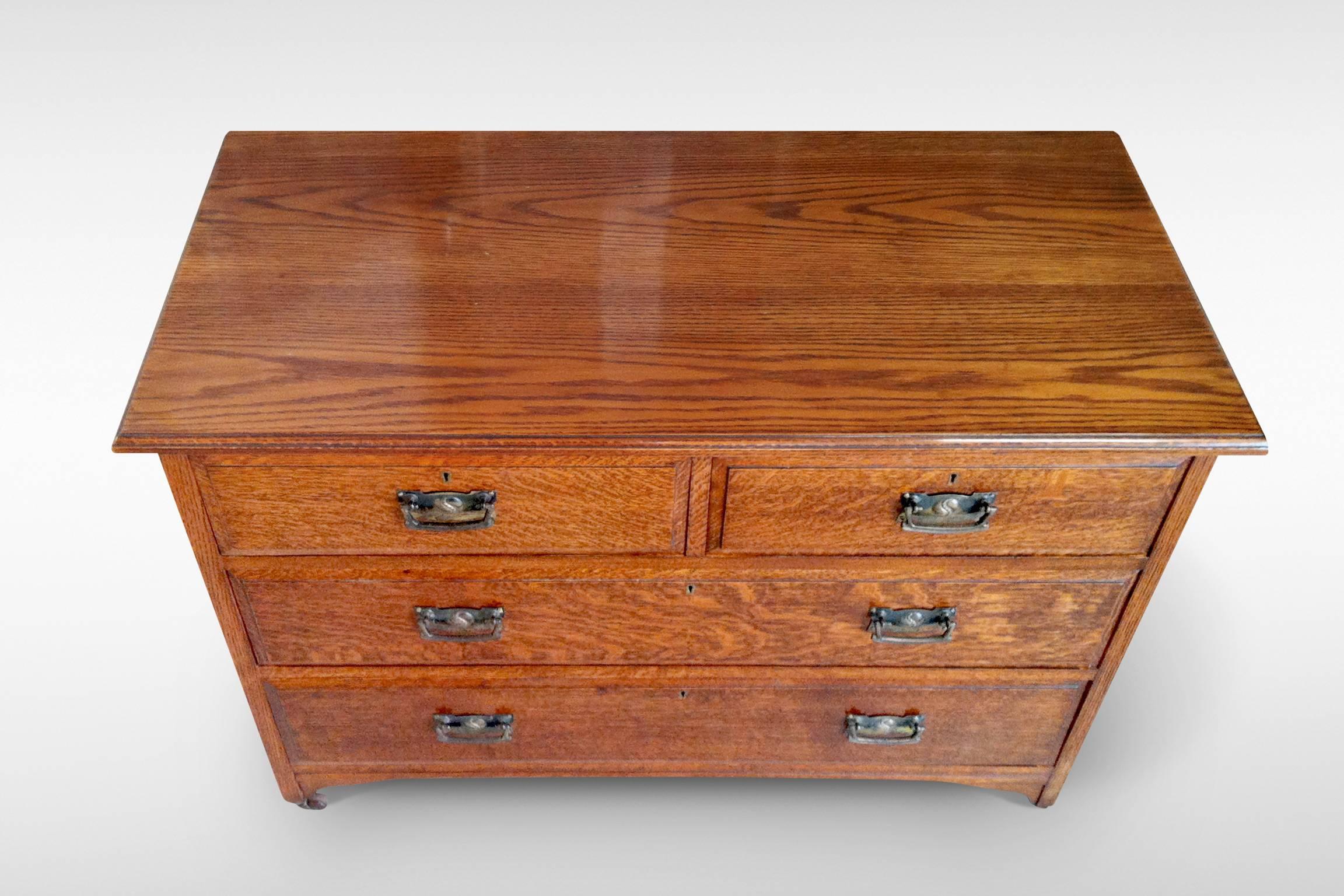 A usefully compact Arts and Crafts chest of drawers in oak, the drawer fronts quarter-sawn with particularly fine figuring. There are two long and two short drawers and the handle plates have a repousse-work motif of stylised pomegranate. The