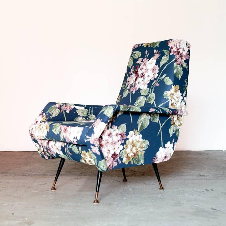 A wooden armchair with metallic feet finished in brass and painted black. It is covered with fabric of beautiful blue floral design, bringing gorgeousness to the space.