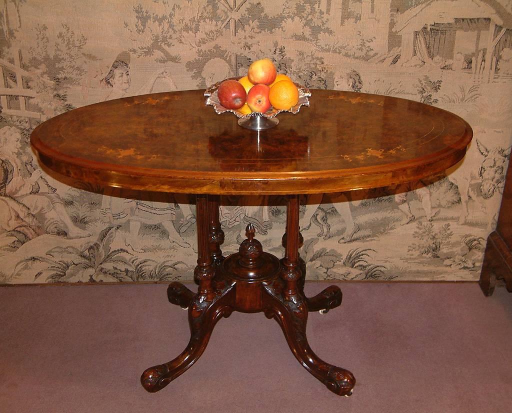 A Victorian figured walnut oval centre table dating from circa 1870. The oval top with Satin wood marquetry inlay above a quatra form base of four grooved columns and standing on four carved cabriole legs terminating with porcelain casters.